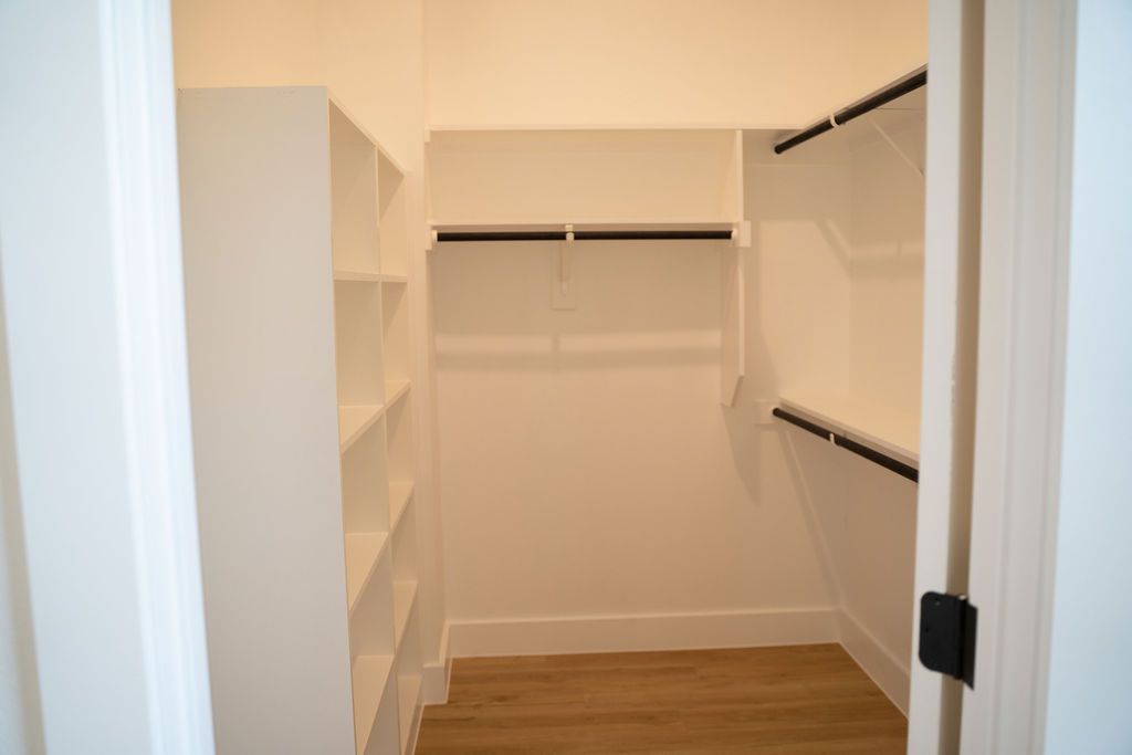 Empty walk-in closet with white walls, shelving on the left, and rods for hanging clothes. Hardwood floor.