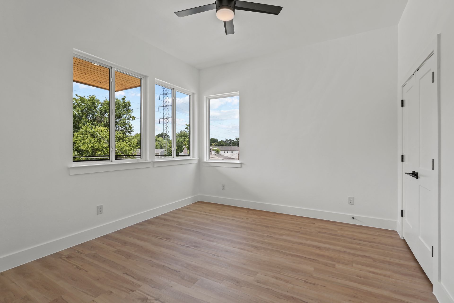 Empty modern bedroom with light wood floors, white walls, and three large windows.