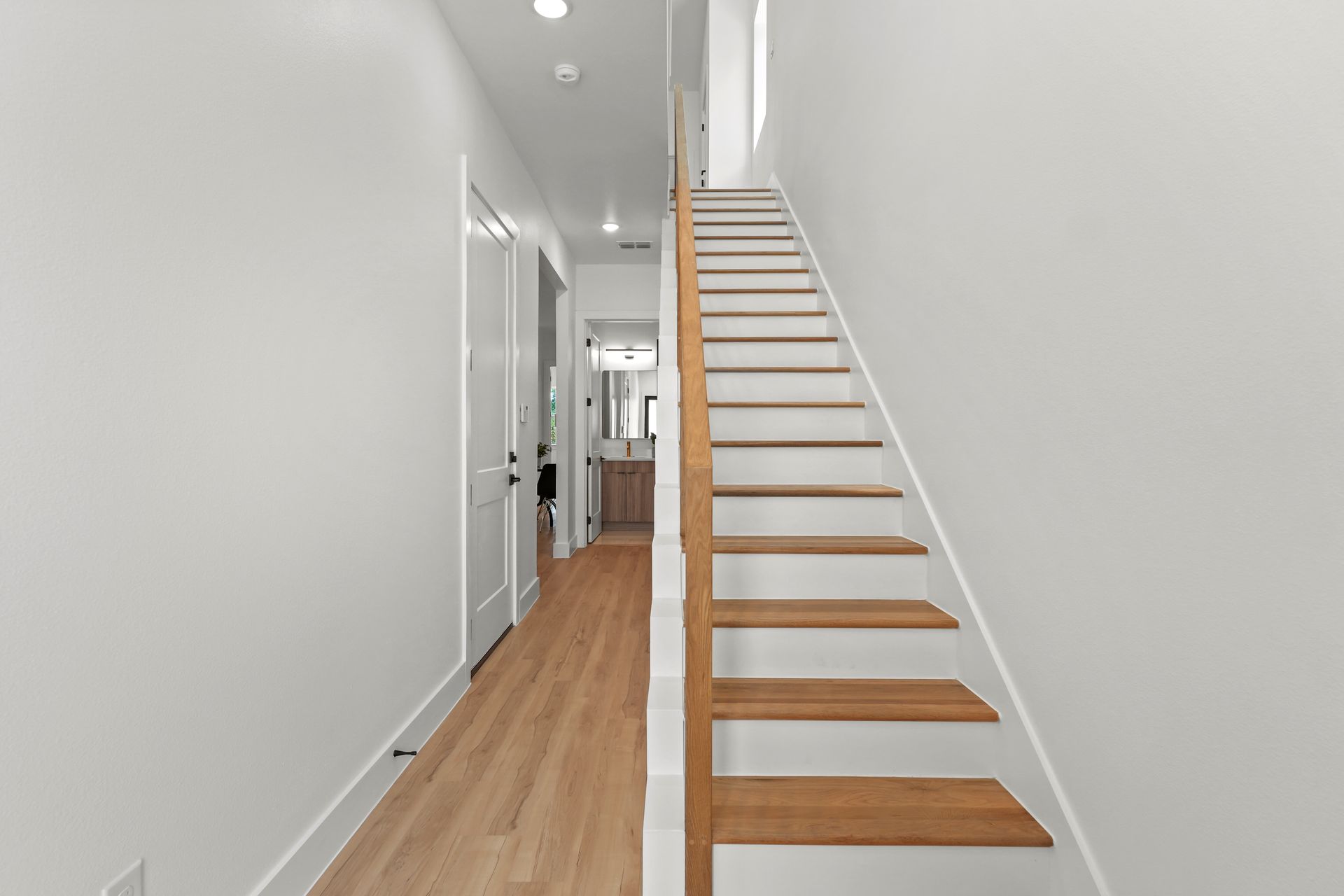 A narrow hallway with wooden floors and stairs leading up to the second floor. White walls and doors line the left side.