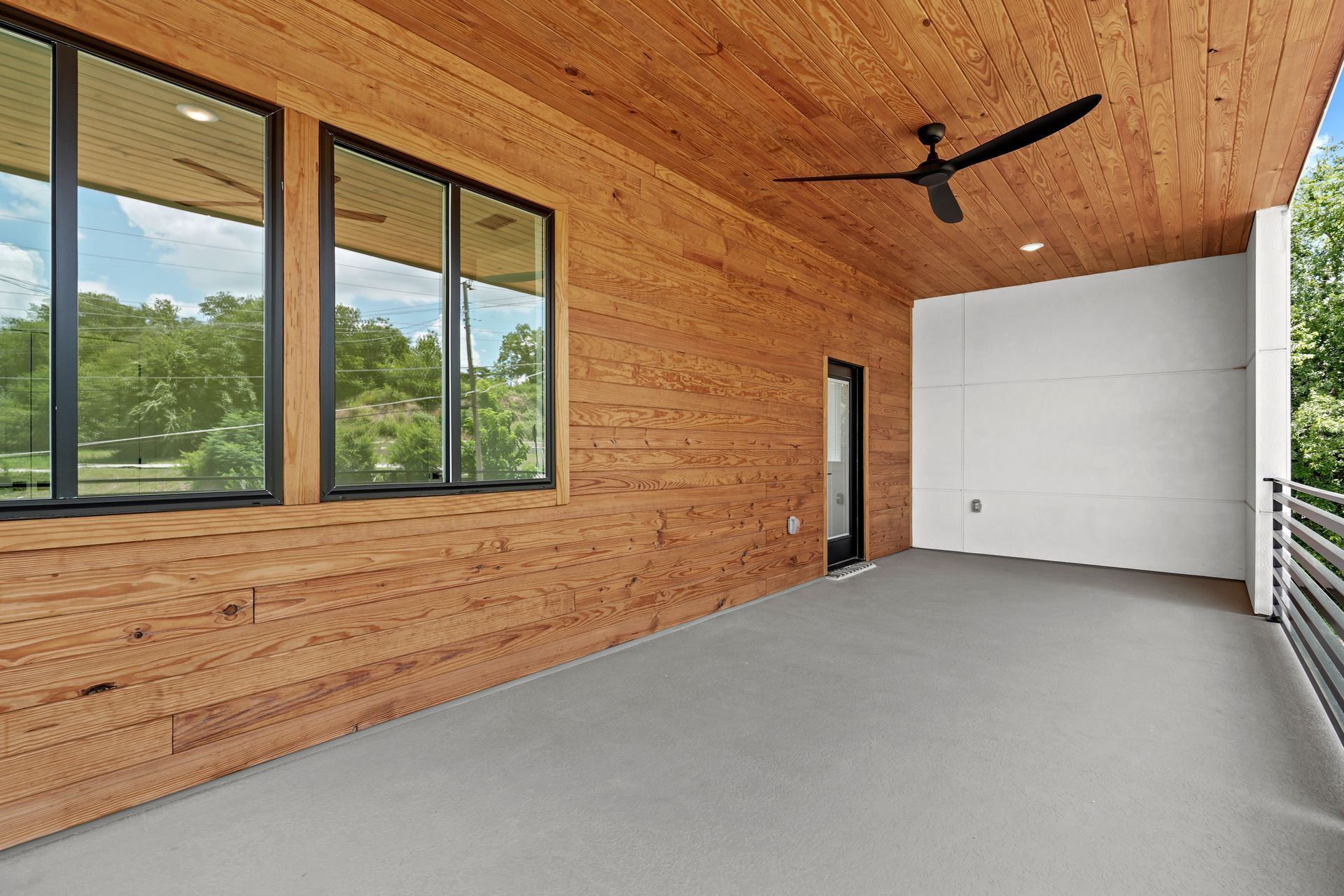 Covered outdoor patio with wood siding and ceiling, gray floor, and black framed windows. A ceiling fan is visible.