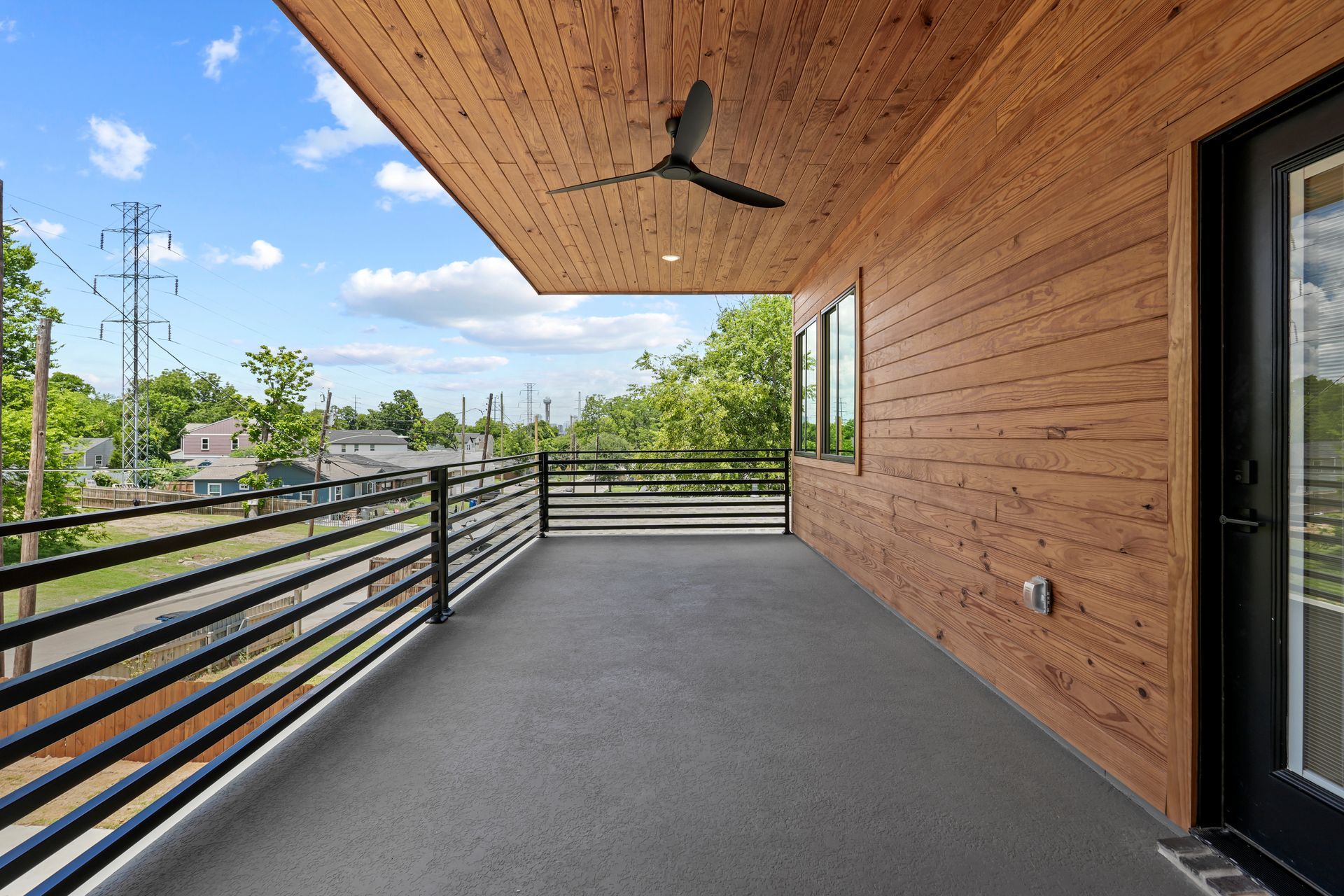 Balcony with a gray floor, wooden ceiling and walls, black railing, and a view of a neighborhood under a blue sky.