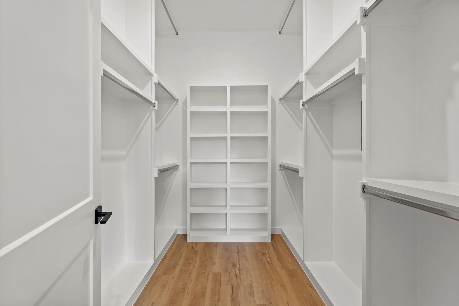 Empty walk-in closet with white shelving, hanging rods, and a central set of shelves against a wooden floor.