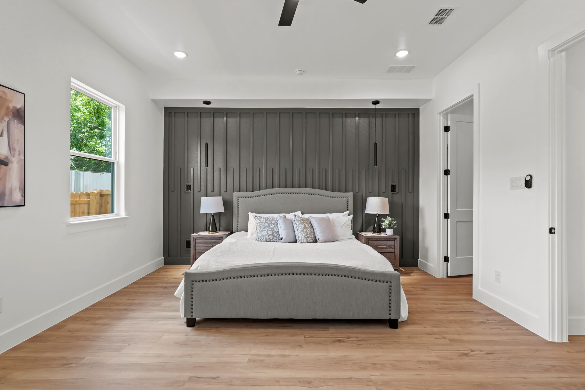 Spacious bedroom with gray upholstered bed against a dark gray paneled accent wall, and light wood floors.