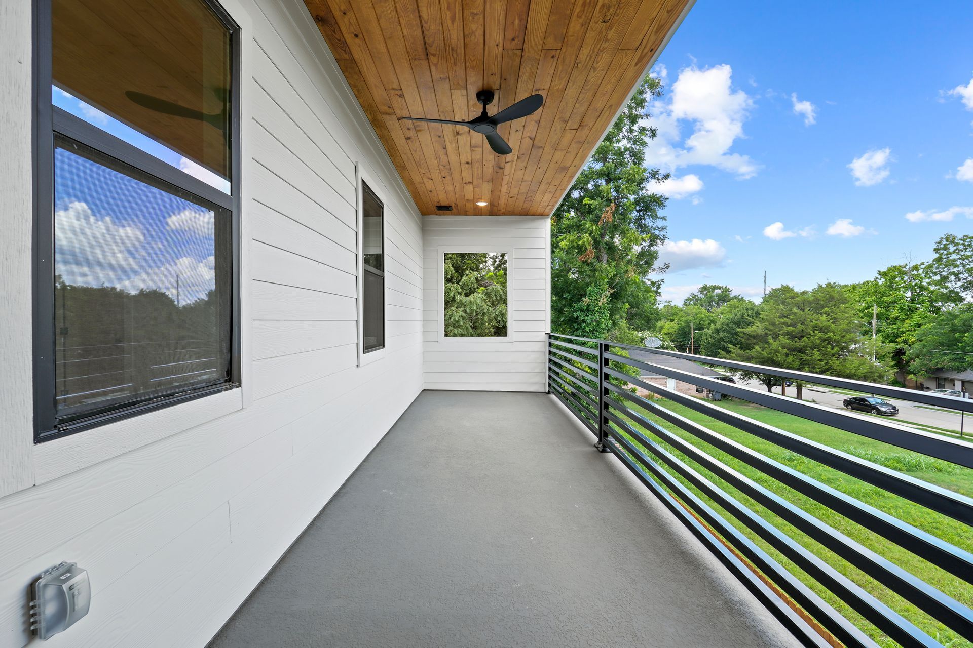 Balcony with a wooden ceiling, white walls, and black railing. View of a street with trees and blue sky.