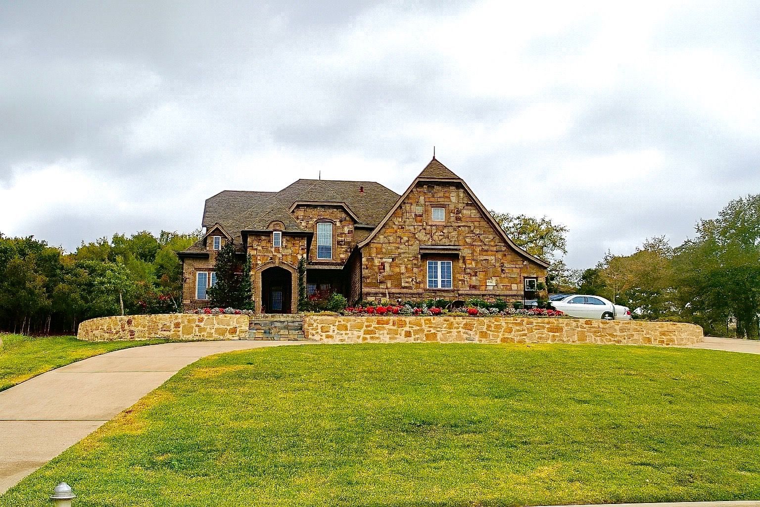 A stone-clad house with a winding walkway and green lawn under a cloudy sky. A car is parked in the driveway.
