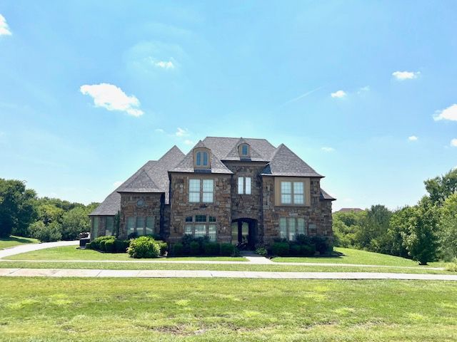 A large, two-story stone house with a dark roof under a blue sky, surrounded by a green lawn and trees.