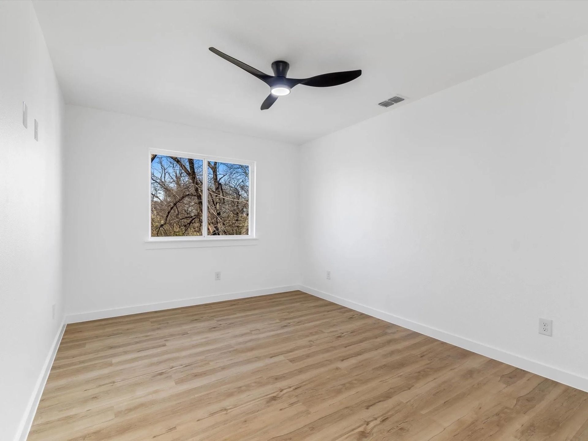Empty, bright room with wood-look flooring and white walls. A window reveals trees outside, and a ceiling fan is mounted.