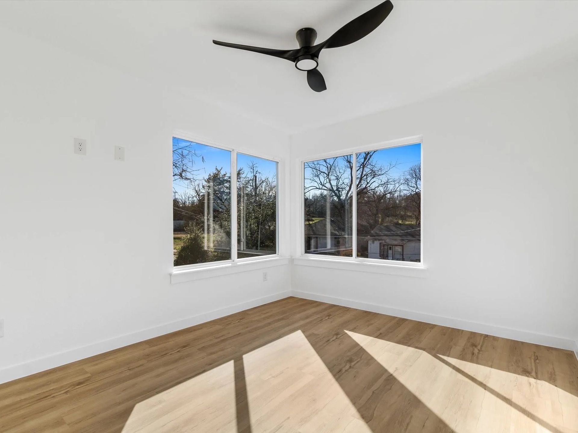 Empty, white room with light wood floors and two windows overlooking a wooded area. A black ceiling fan is visible.