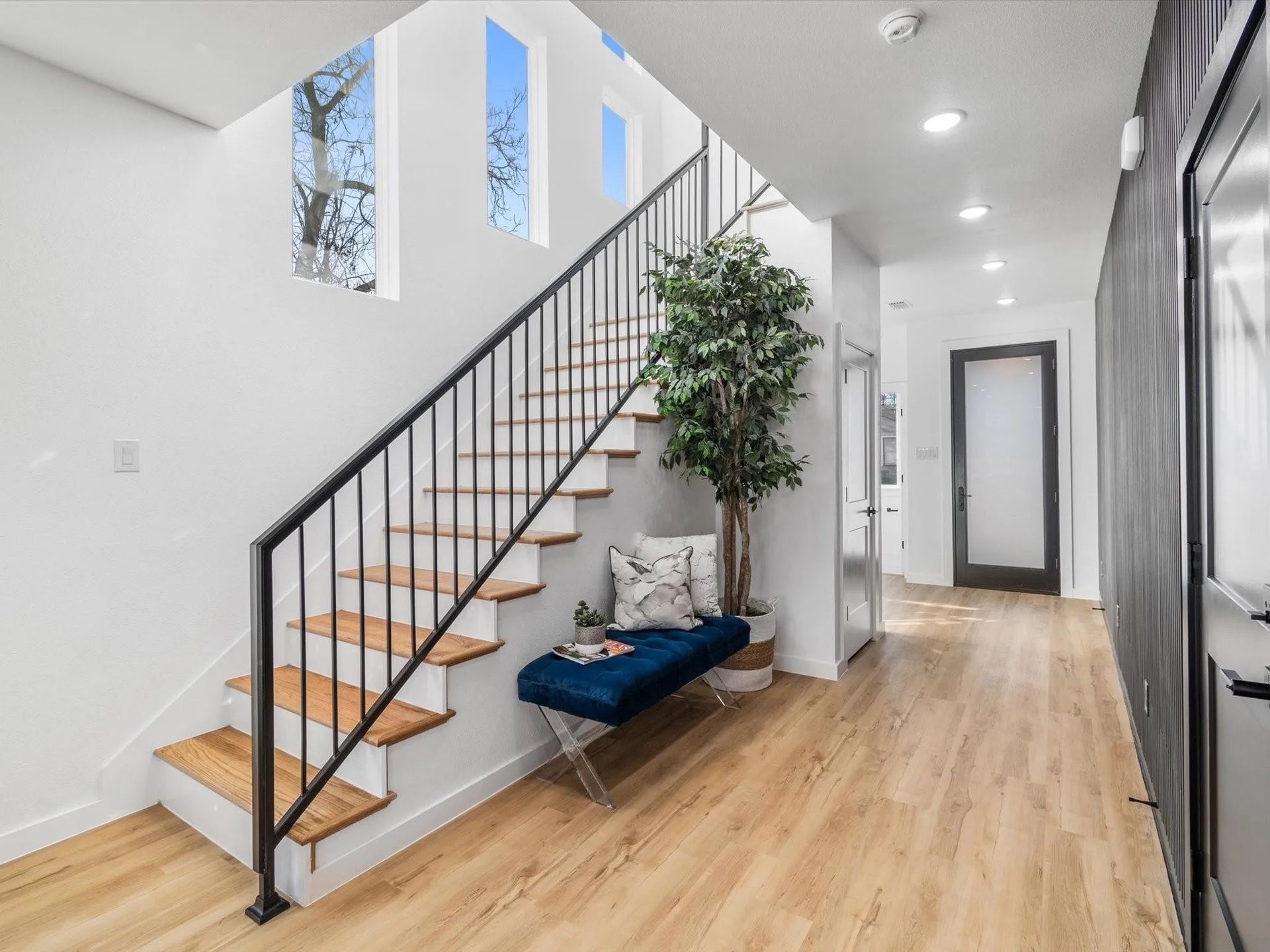 Interior hallway with wooden floors, a staircase with black railing, and a blue bench with a plant.