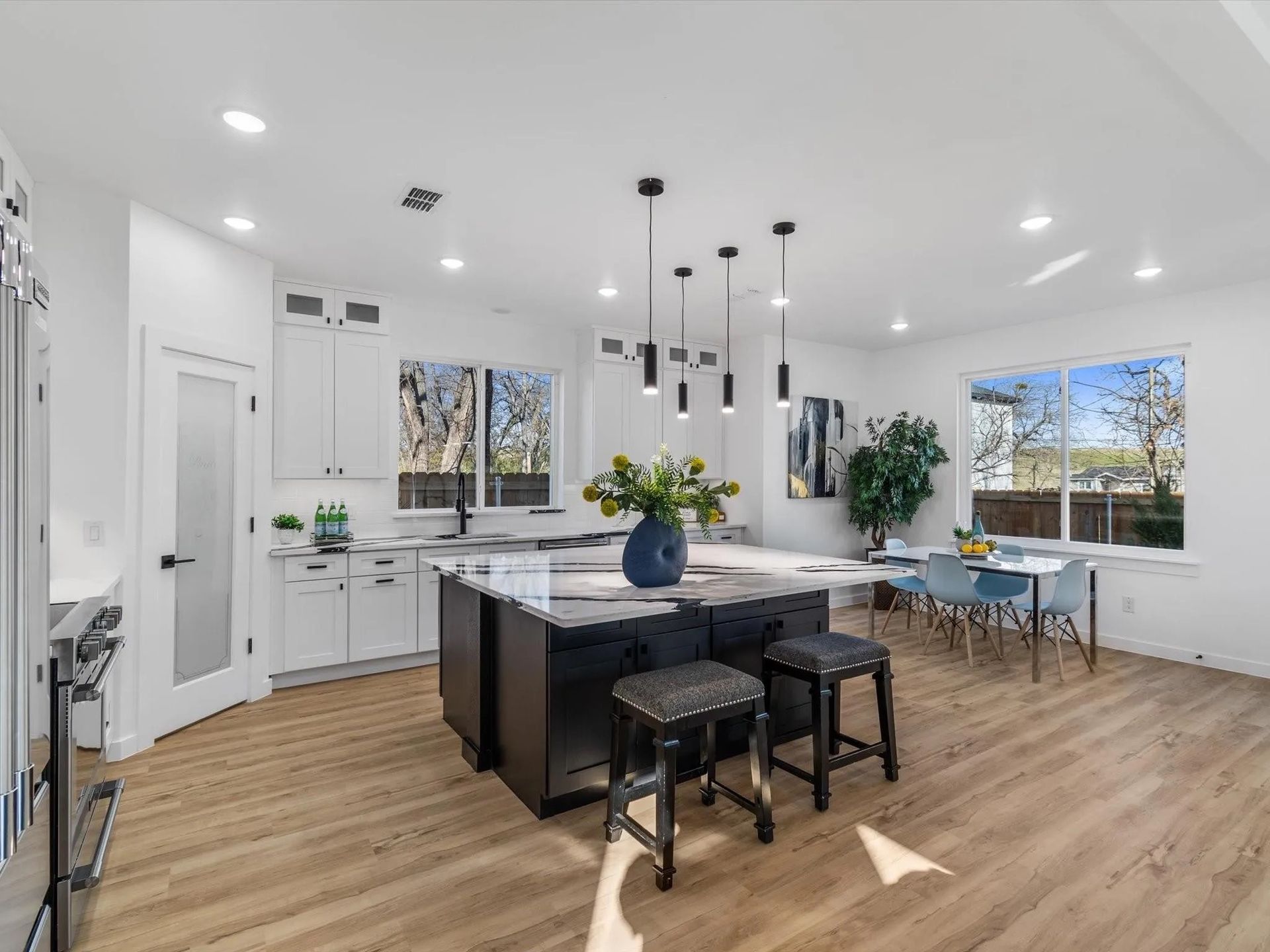 Modern kitchen with dark island, white cabinets, and dining area by a window. Light wood flooring and overhead lighting.