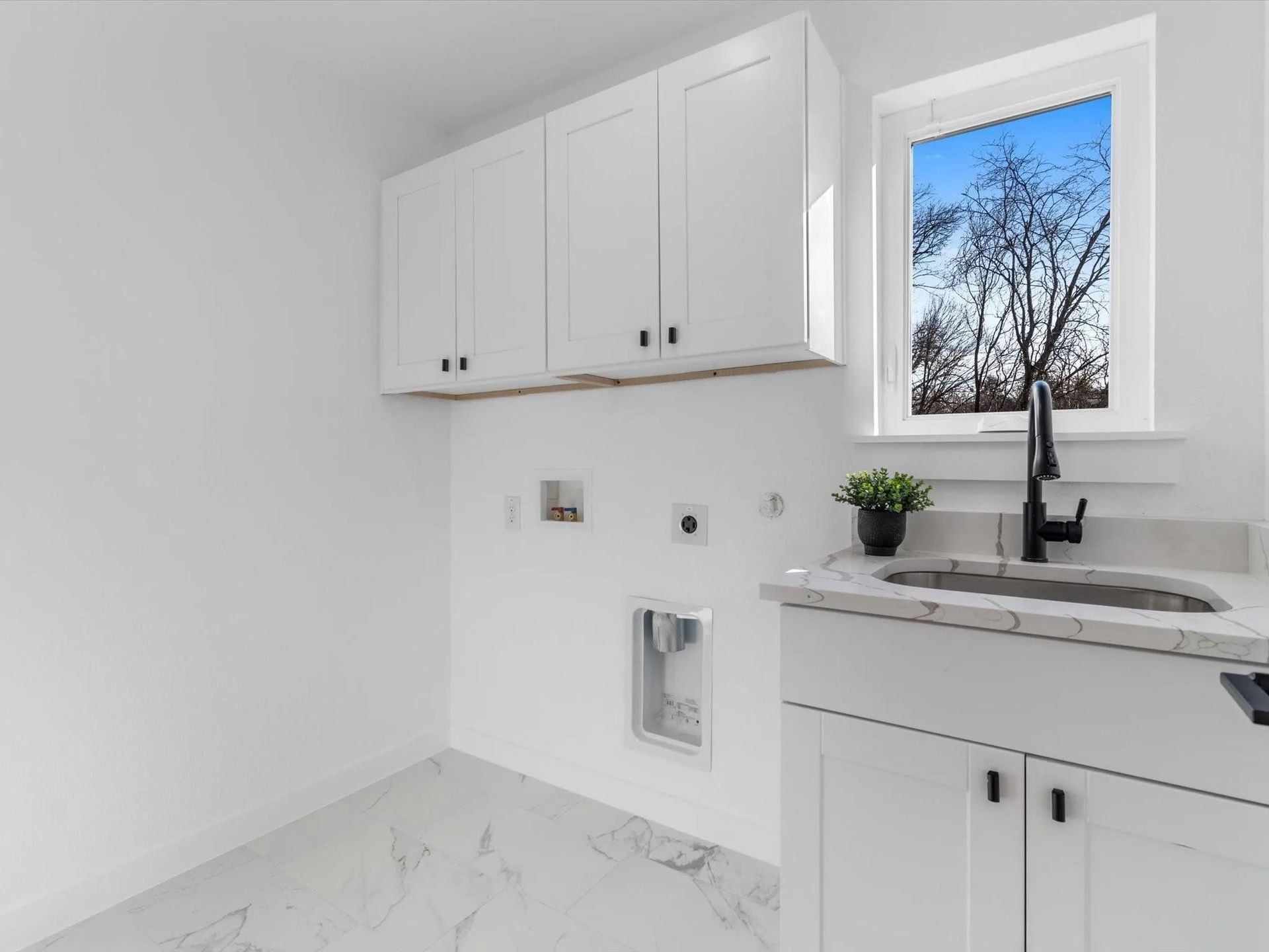 A bright white laundry room with a sink, cabinetry, and a window with a view of a tree.