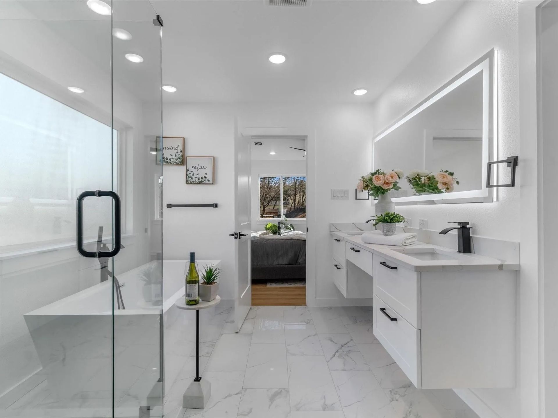 Modern white bathroom with marble floors, glass shower, and floating vanity. The doorway opens to a glimpse of a bedroom.
