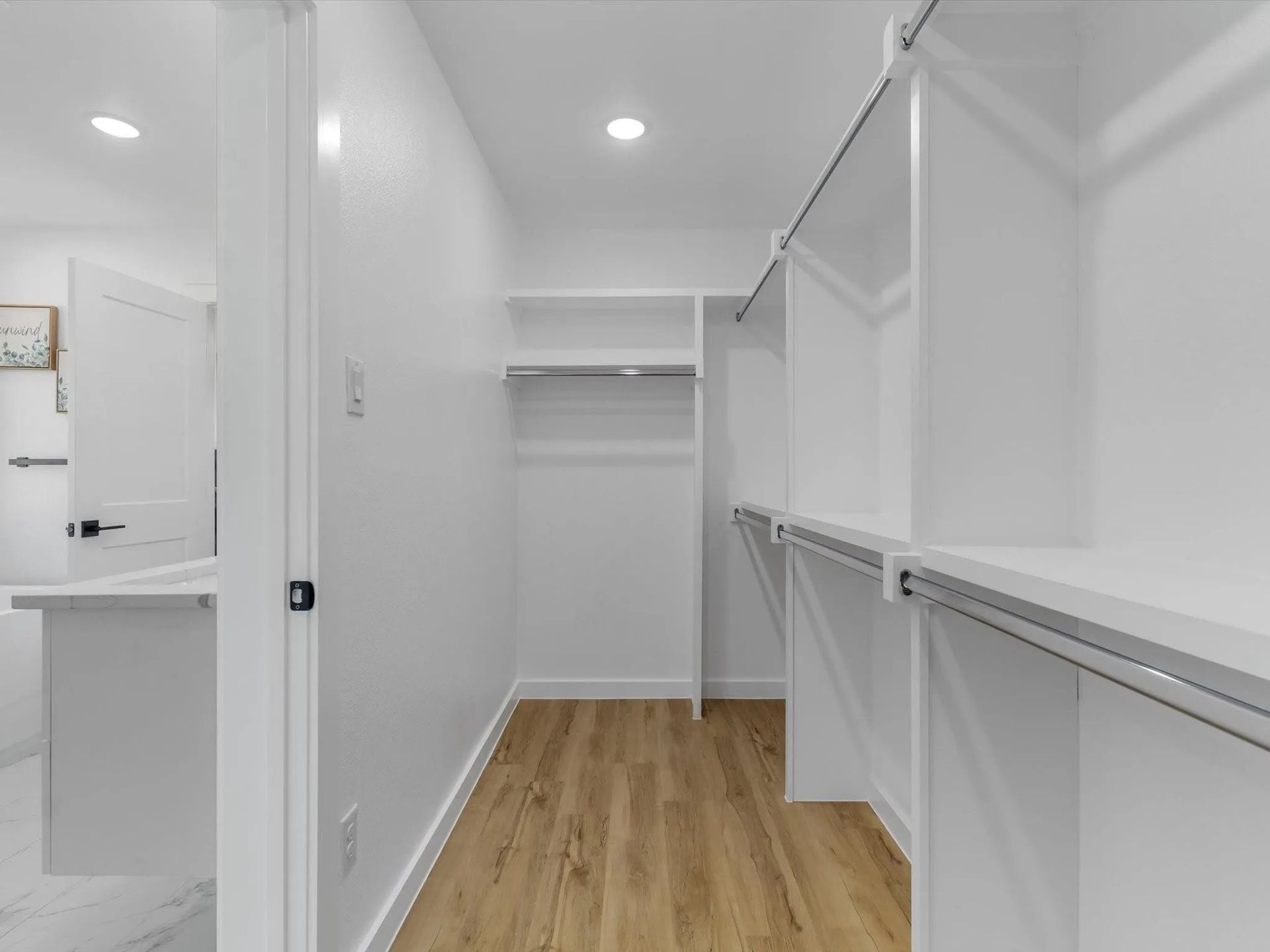An empty, bright white walk-in closet with wooden floors and built-in shelving and rods. 