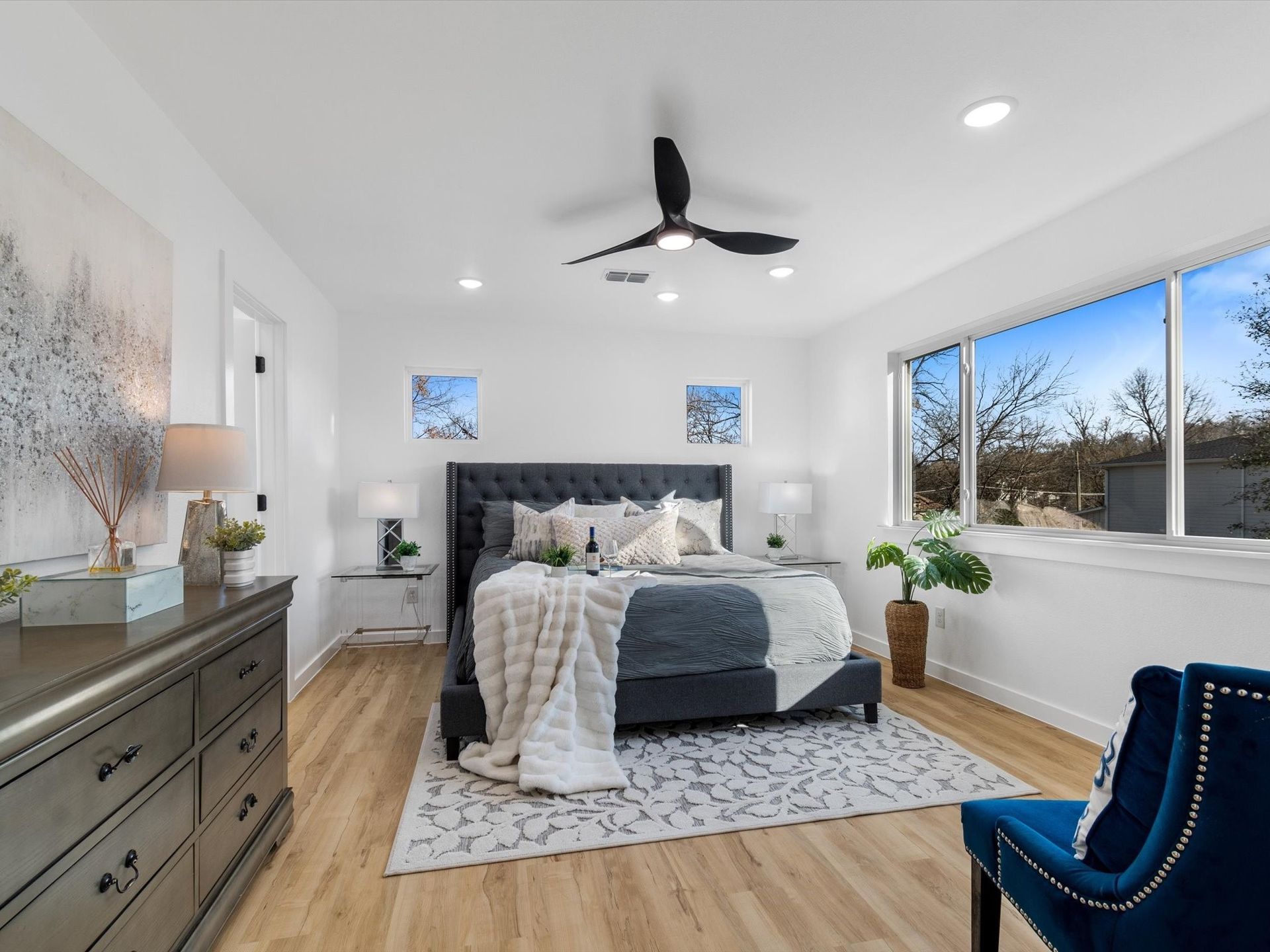 Spacious bedroom with a king-sized bed, dresser, and a blue velvet chair, with a view through a large window.