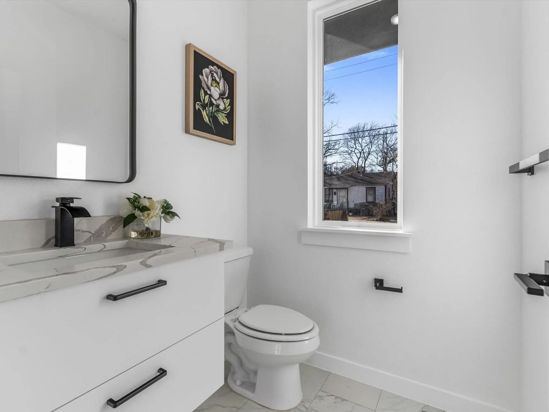 Bright white bathroom with a vanity, toilet, and window overlooking a cityscape. A floral painting hangs on the wall.