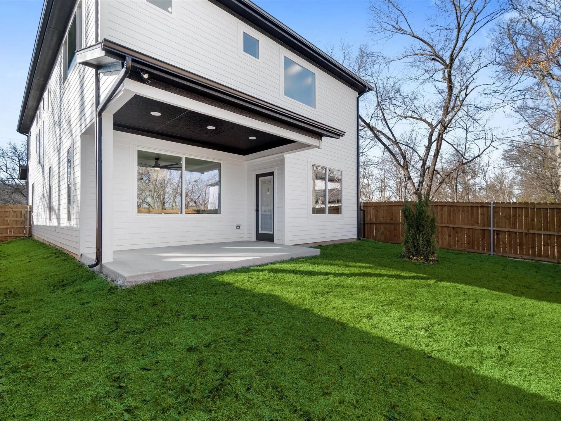 Backyard view of a modern two-story house with a covered patio, surrounded by a green lawn and a wooden fence.