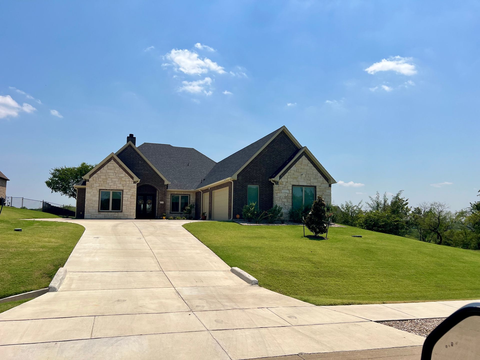 Beige stone house with a dark roof and a long concrete driveway on a sunny day with a blue sky.