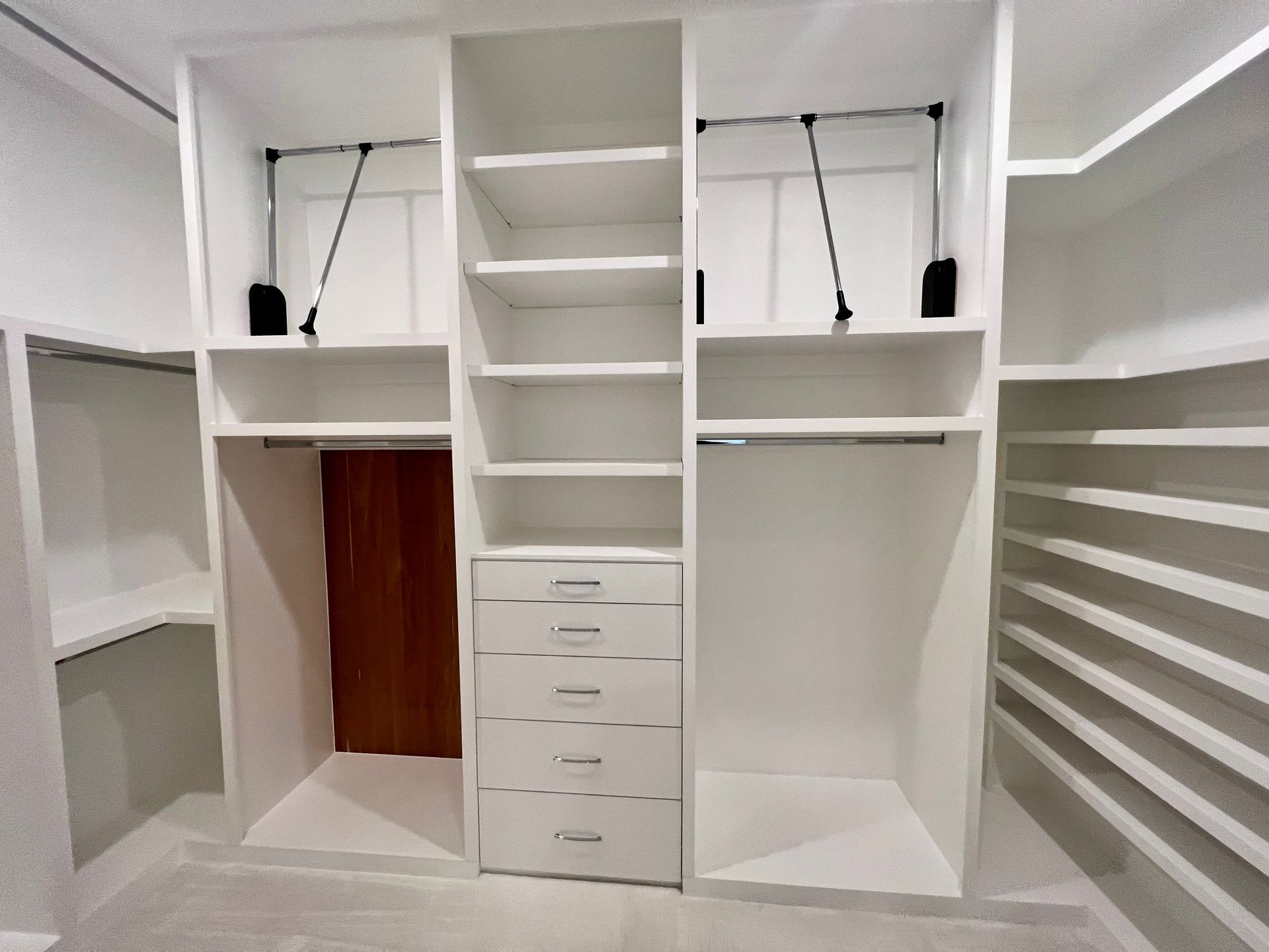 White closet with multiple shelves, drawers, and hanging rods. There's a small door and open space for hanging clothes.
