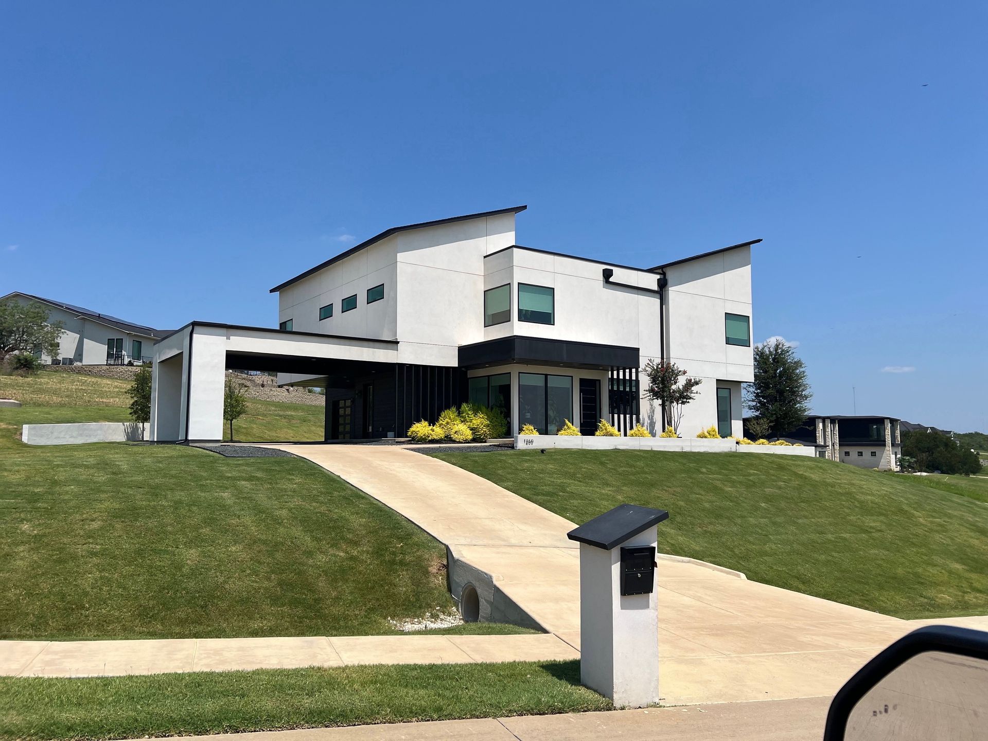Modern white house with black trim, a sloping driveway, and a grassy lawn under a clear blue sky.