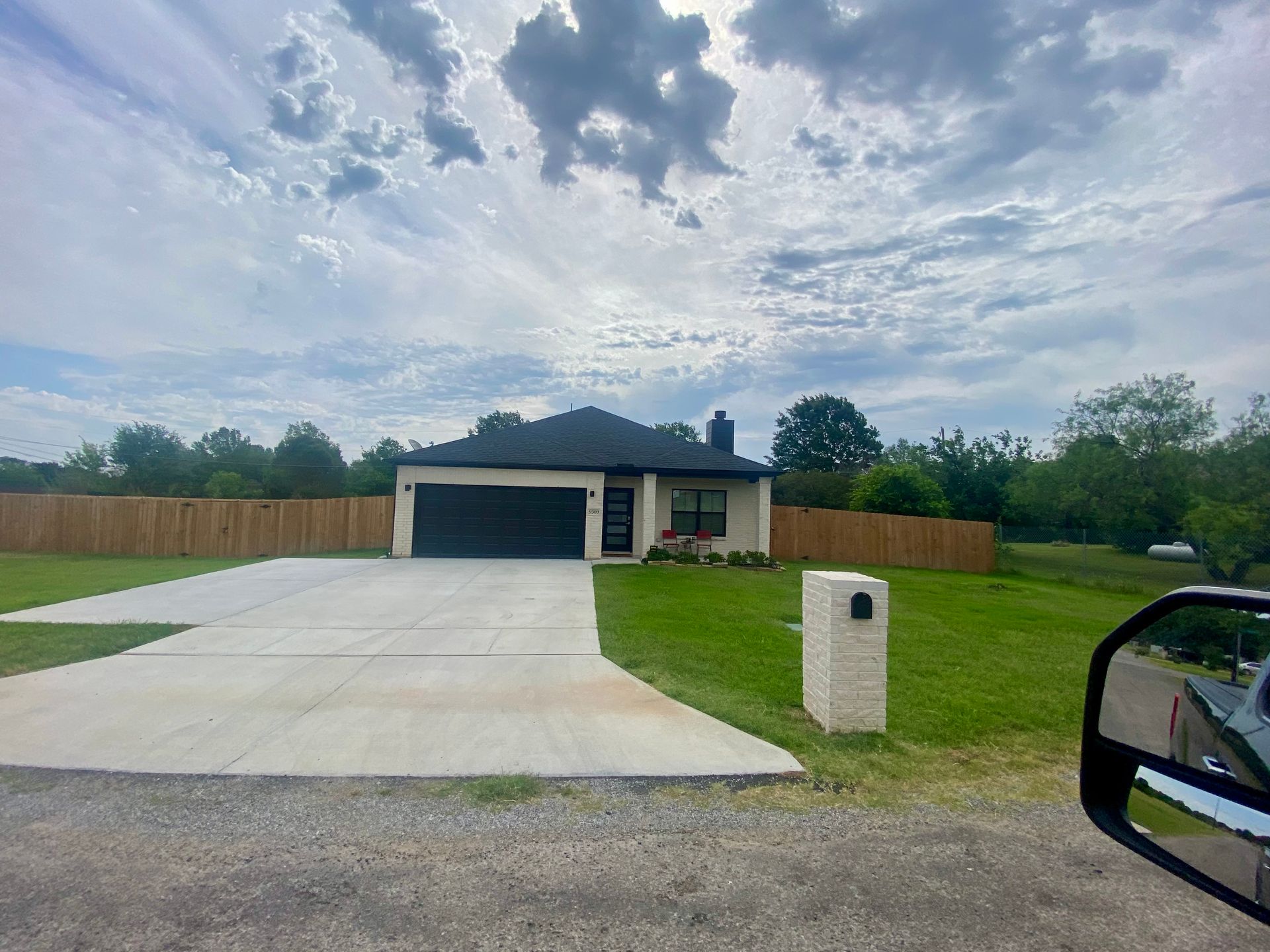 A single-story house with a black garage door, surrounded by a wooden fence and green lawn, under a cloudy sky.