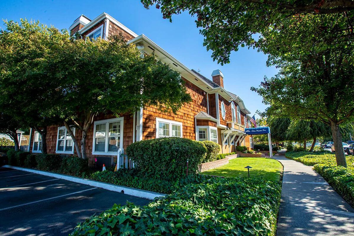 Two-story brown shingle building with trees and landscaping under a blue sky.
