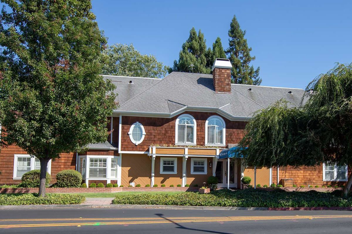Two-story house with brown shingle siding, tan walls, gray roof, and arched windows. Trees flank sides.