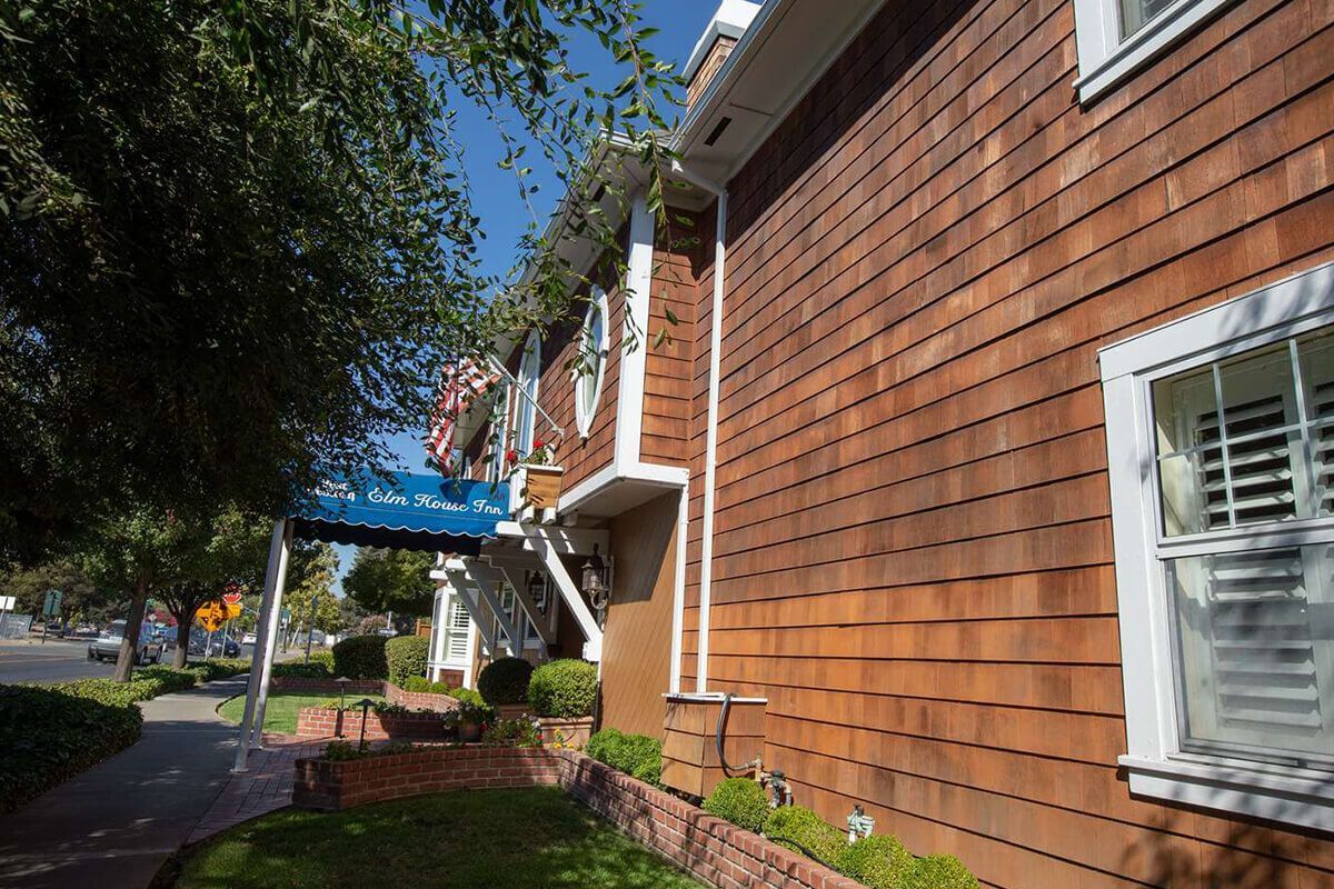 Brown shingle building with white trim, blue awning, and sidewalk alongside a road.