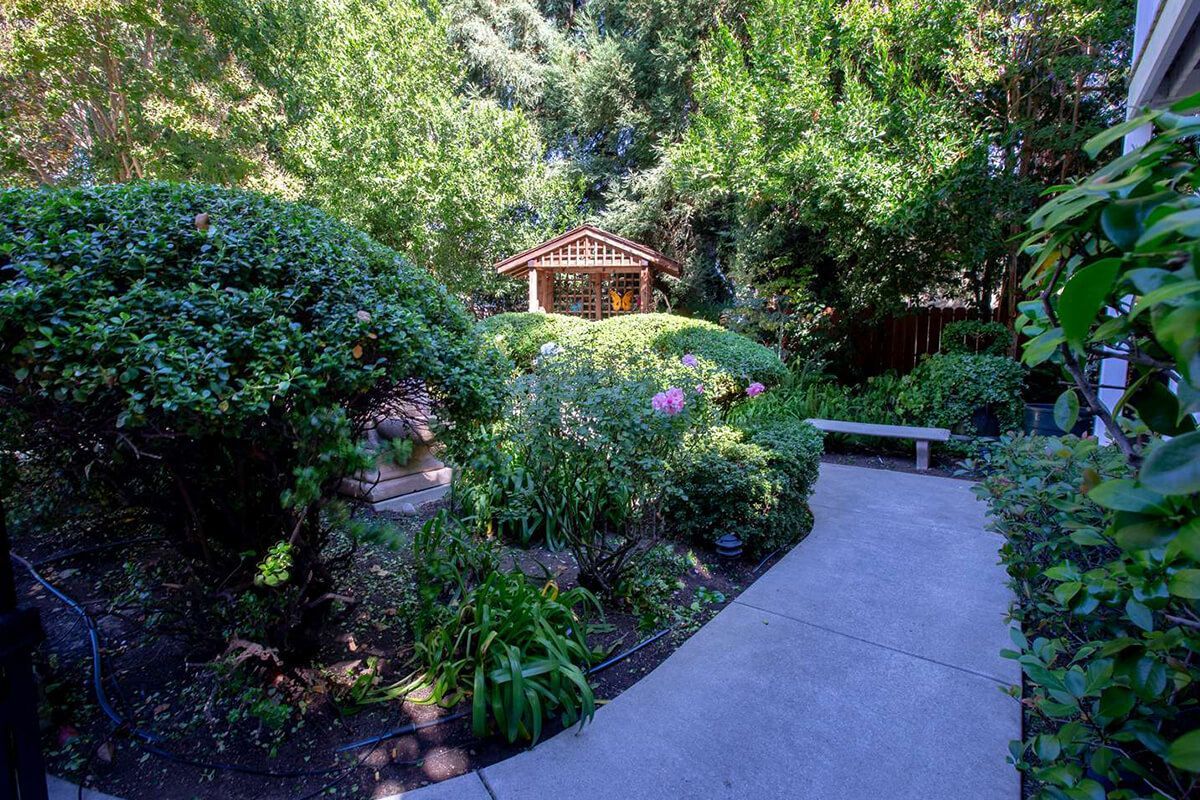 Pathway through a lush garden with a gazebo in the background. Green foliage, stone bench. Sunny day.