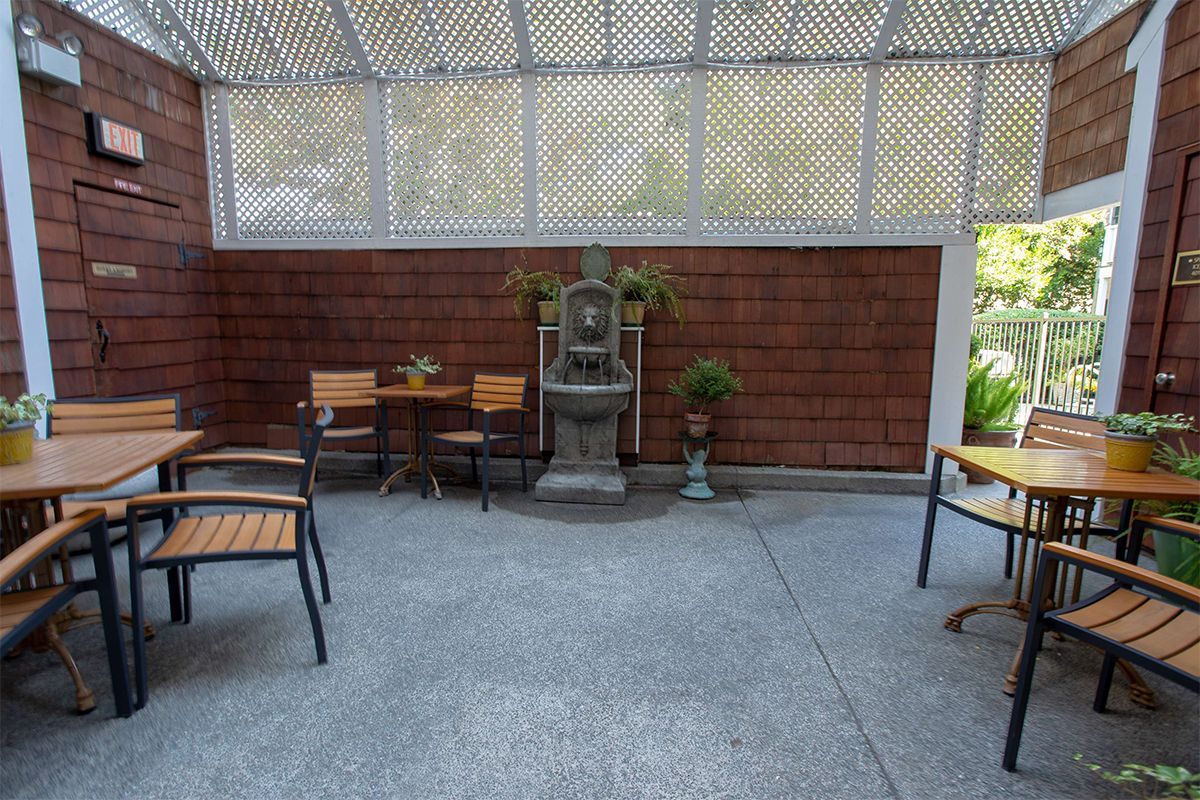 Outdoor dining area with tables, chairs, and a fountain. Brown brick walls, textured roof, grey floor.