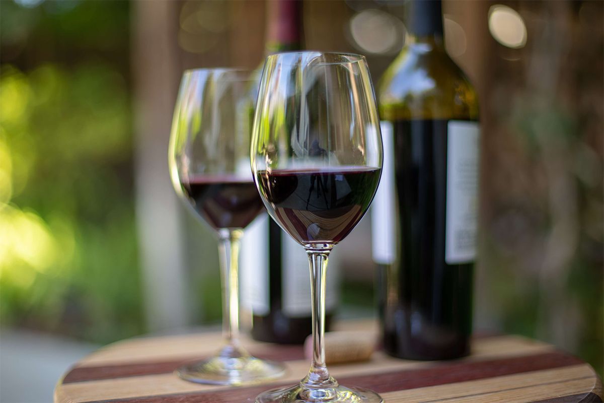 Two wine glasses filled with red wine, behind two wine bottles on a wooden surface.