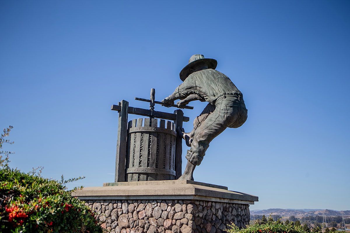 Bronze statue of a person pressing grapes, located outdoors with a clear blue sky background.