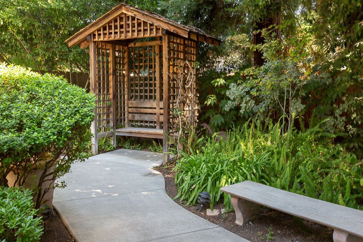Stone path leads to wooden gazebo in a lush garden with a bench on the right.