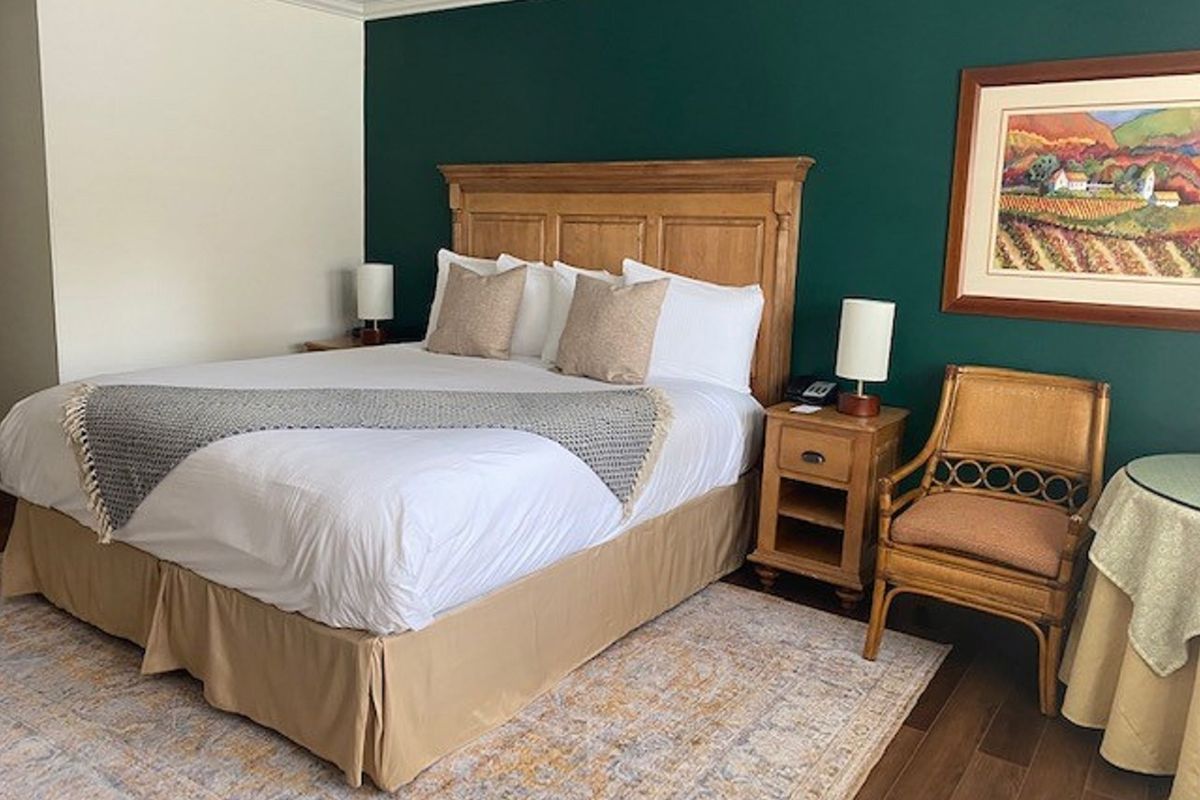 Bedroom with a large bed, nightstand, wicker chair, and painting on a teal wall.