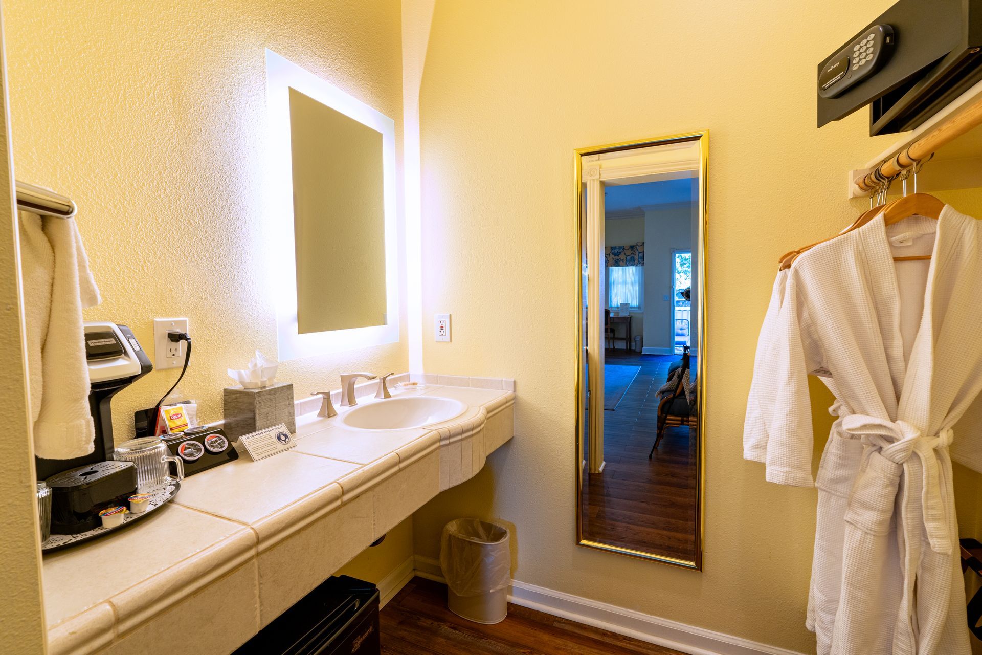 Bathroom with a lit mirror above a sink, towel rack with robe, and a mirror reflecting a room.