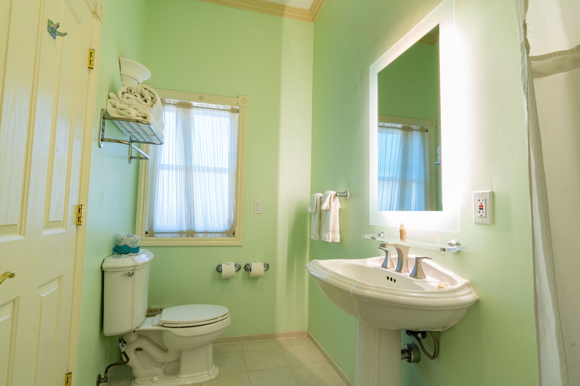 Green-walled bathroom with toilet, sink, mirror, and window with sheer curtains. Towels and toiletries visible.