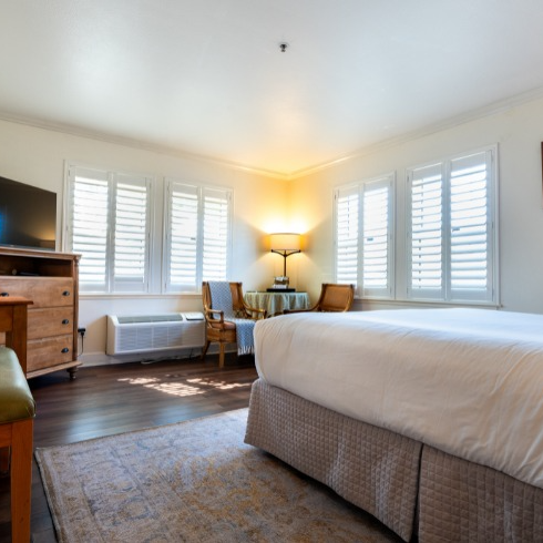 Hotel room with bed, windows with shutters, dresser, rug, and chairs.