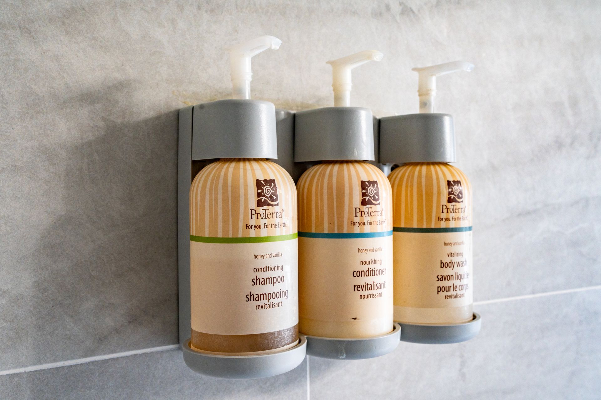 Three beige toiletries bottles mounted on a gray wall, labeled shampoo, conditioner, and body wash.