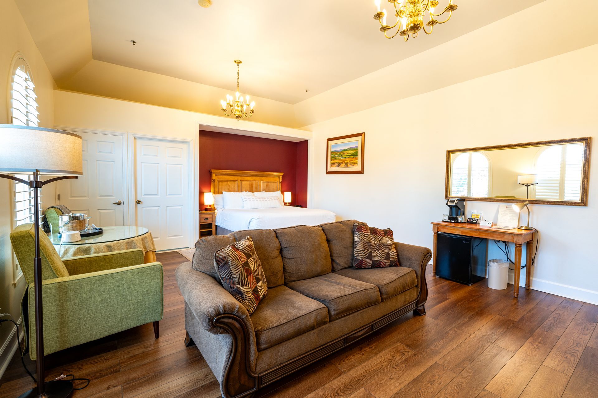 Hotel room with a bed, sofa, armchair, and desk; wooden floors, cream walls, and chandeliers.