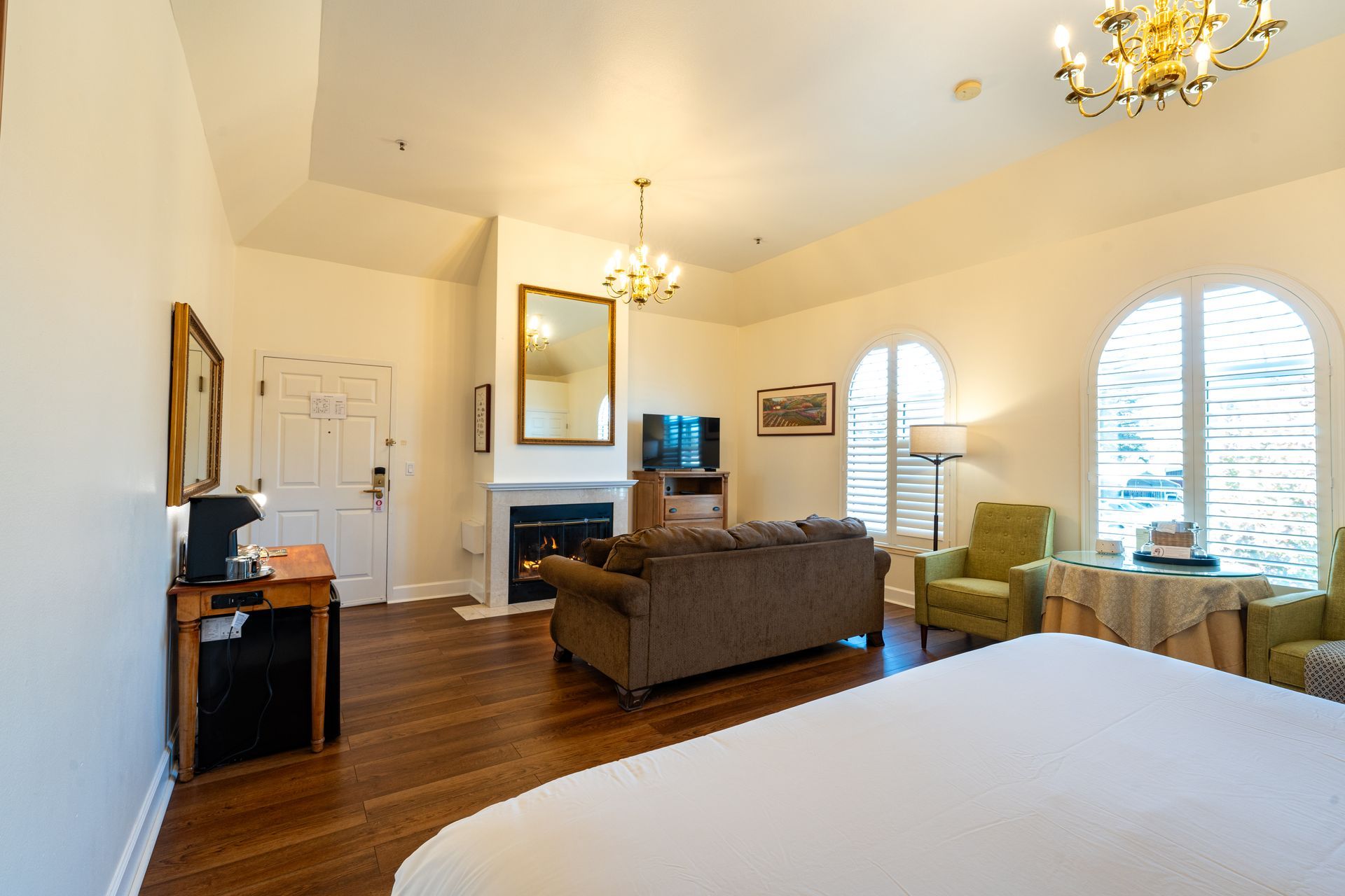 Spacious hotel room with a fireplace, sofa, and a large bed, featuring arched windows and chandeliers.