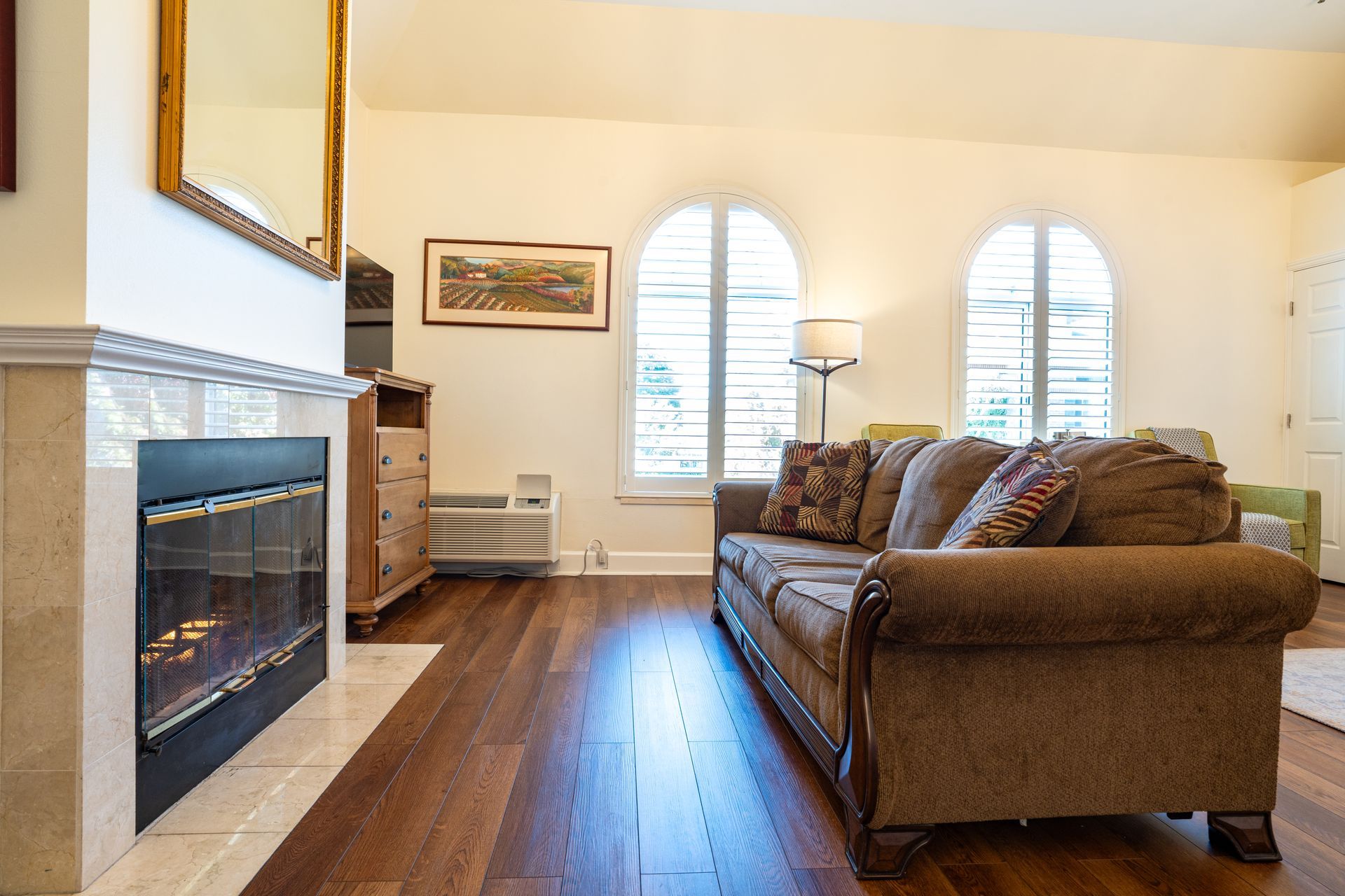 Living room with fireplace, brown sofa, arched windows with shutters, and hardwood floors.
