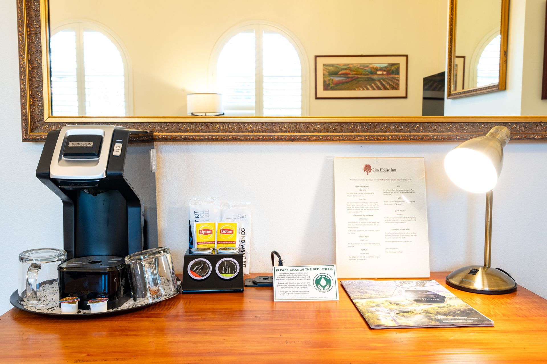 Coffee station on a desk with a Keurig machine, mugs, supplies, lamp, and framed mirror.