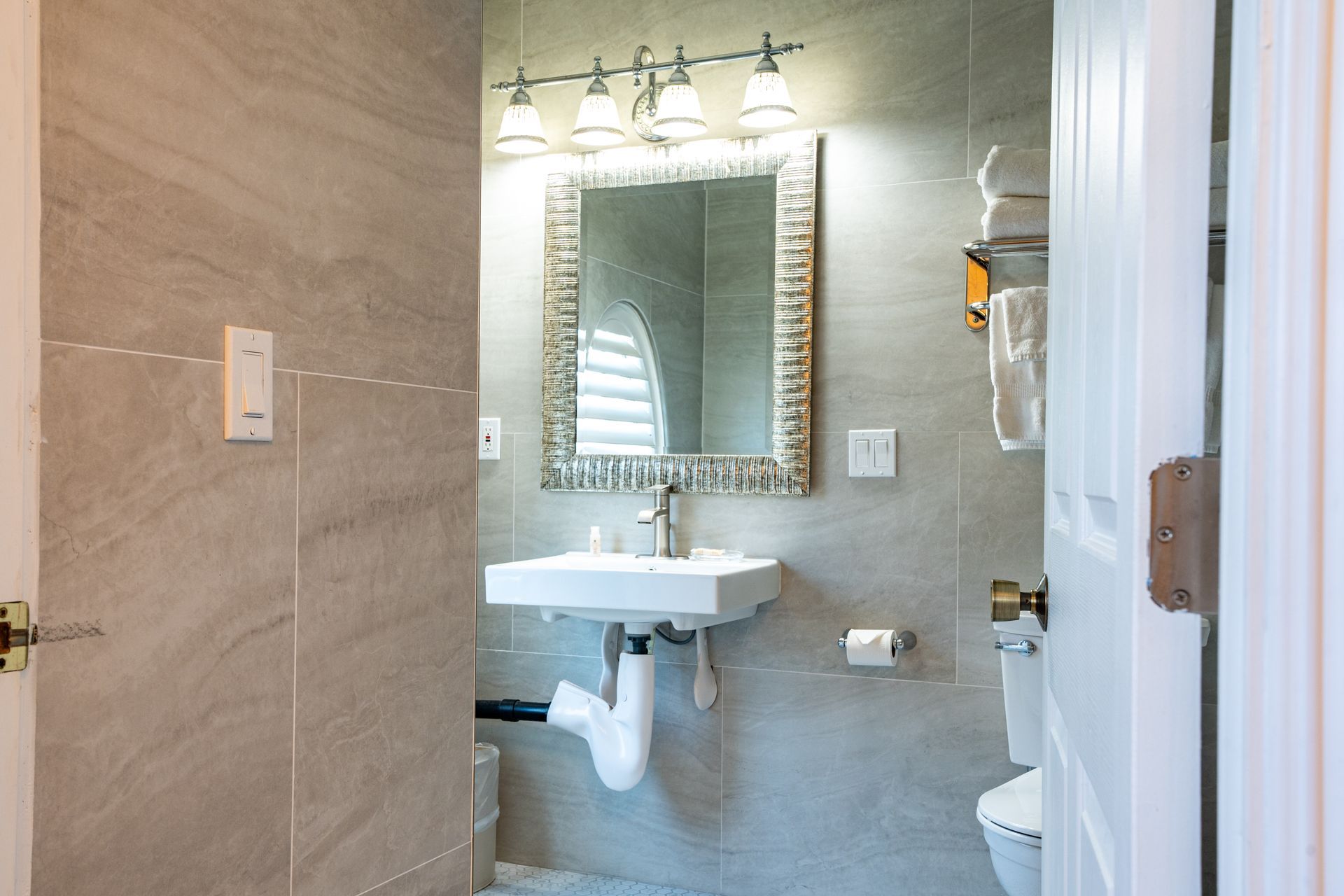 Bathroom with a white sink, toilet, and towels. Gray tiled walls with a mirror and lighting.