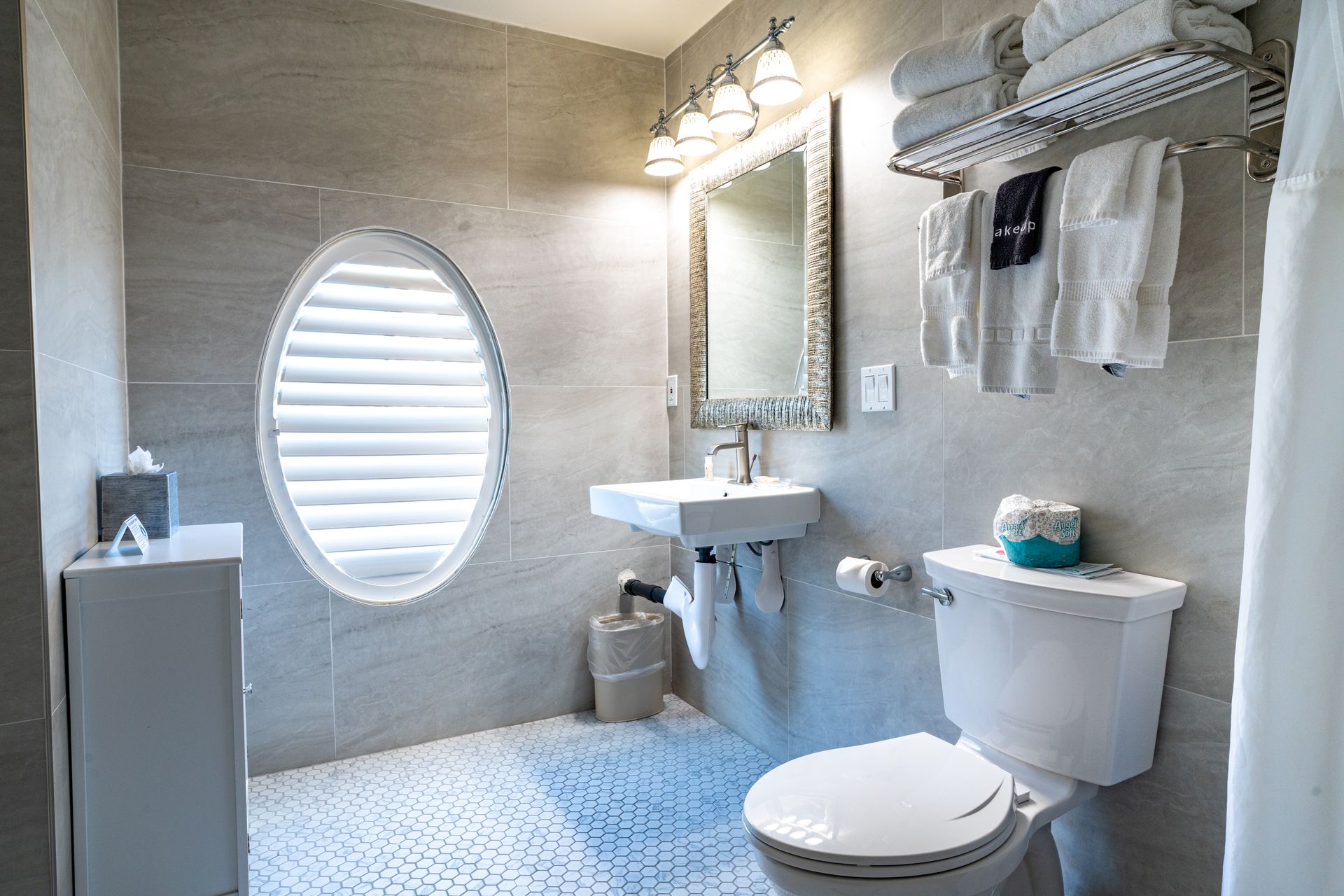 Bathroom with gray tiled walls, an oval window, and white fixtures. Towel rack with folded towels.