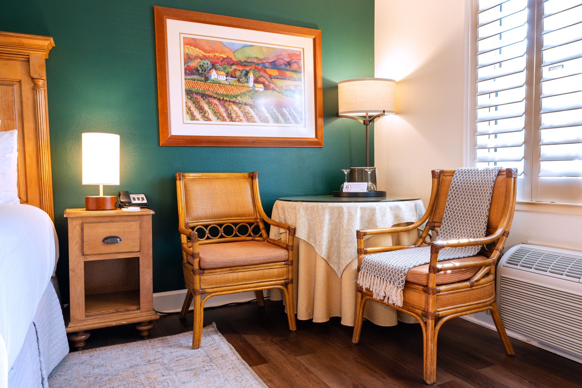 Cozy hotel room with two wicker chairs, a small table, and landscape art on a teal wall.