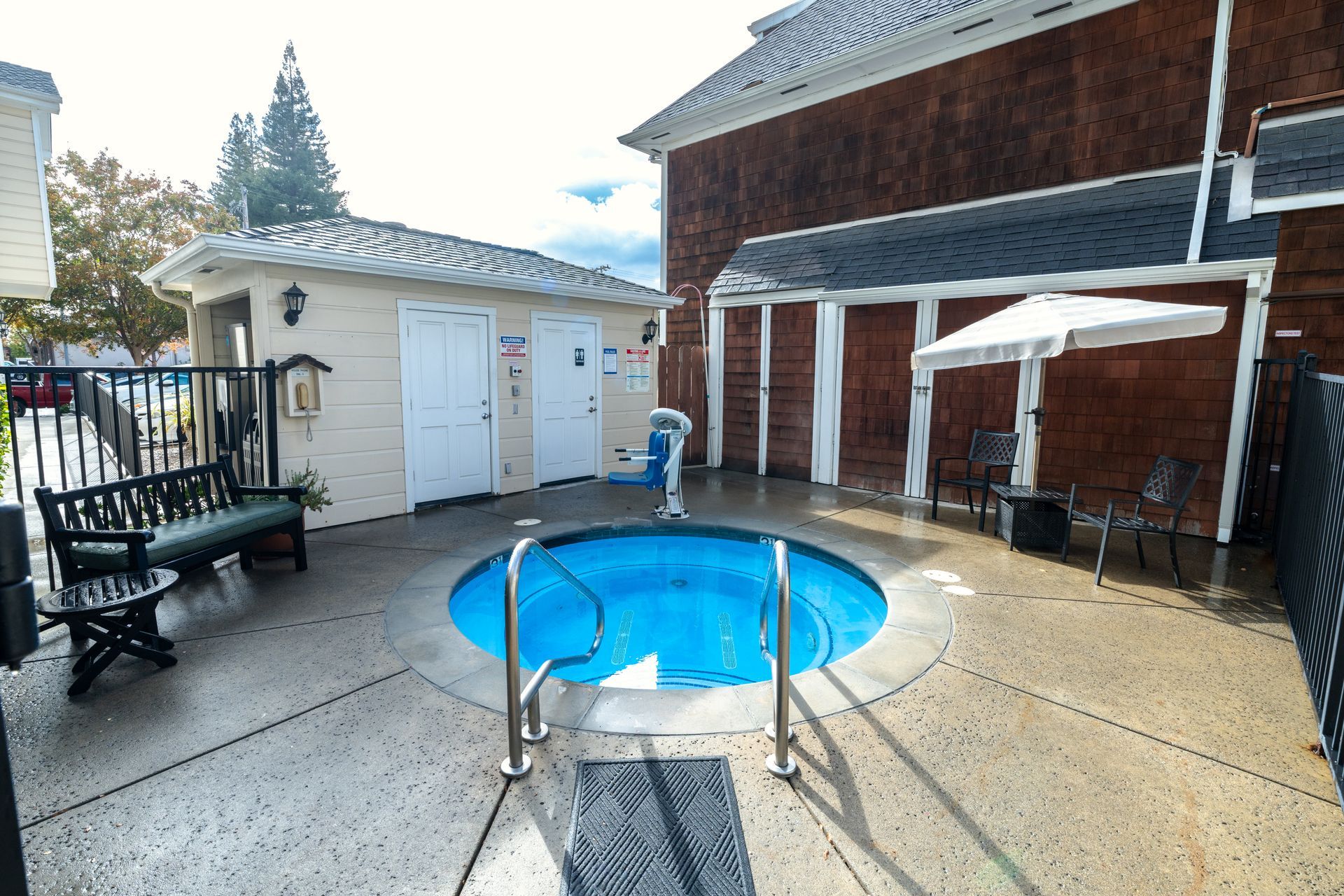 Hot tub surrounded by concrete patio, near a small building and a brown building.