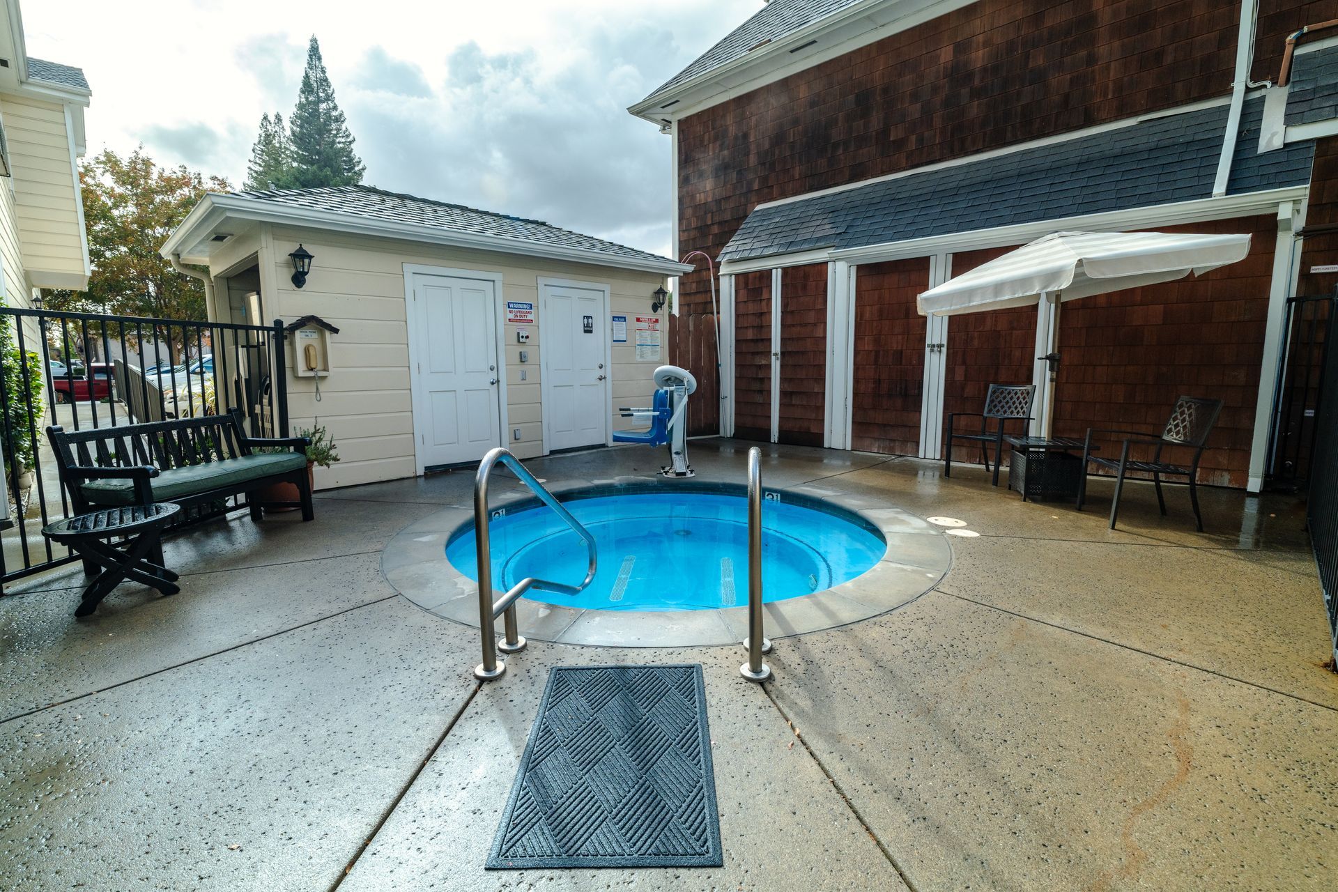 Outdoor hot tub surrounded by concrete, with a changing room building and seating. Overcast sky.