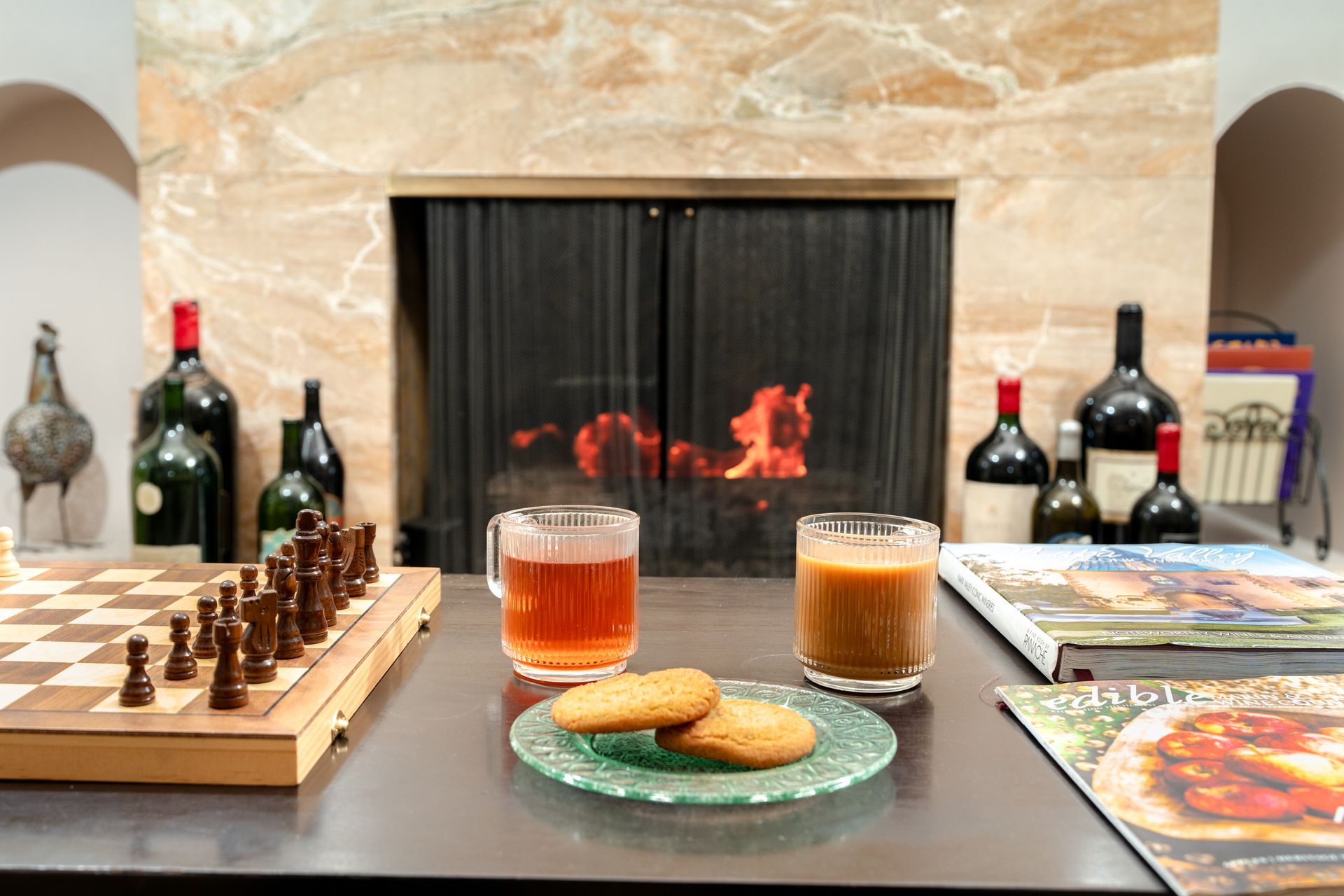 Cozy scene with a fireplace, drinks, cookies, chess set, and wine bottles.