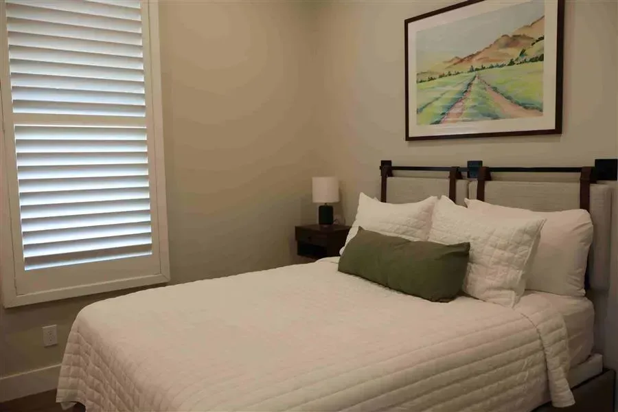 Bedroom with a white bed, shutters, art, and a small lamp on a nightstand.