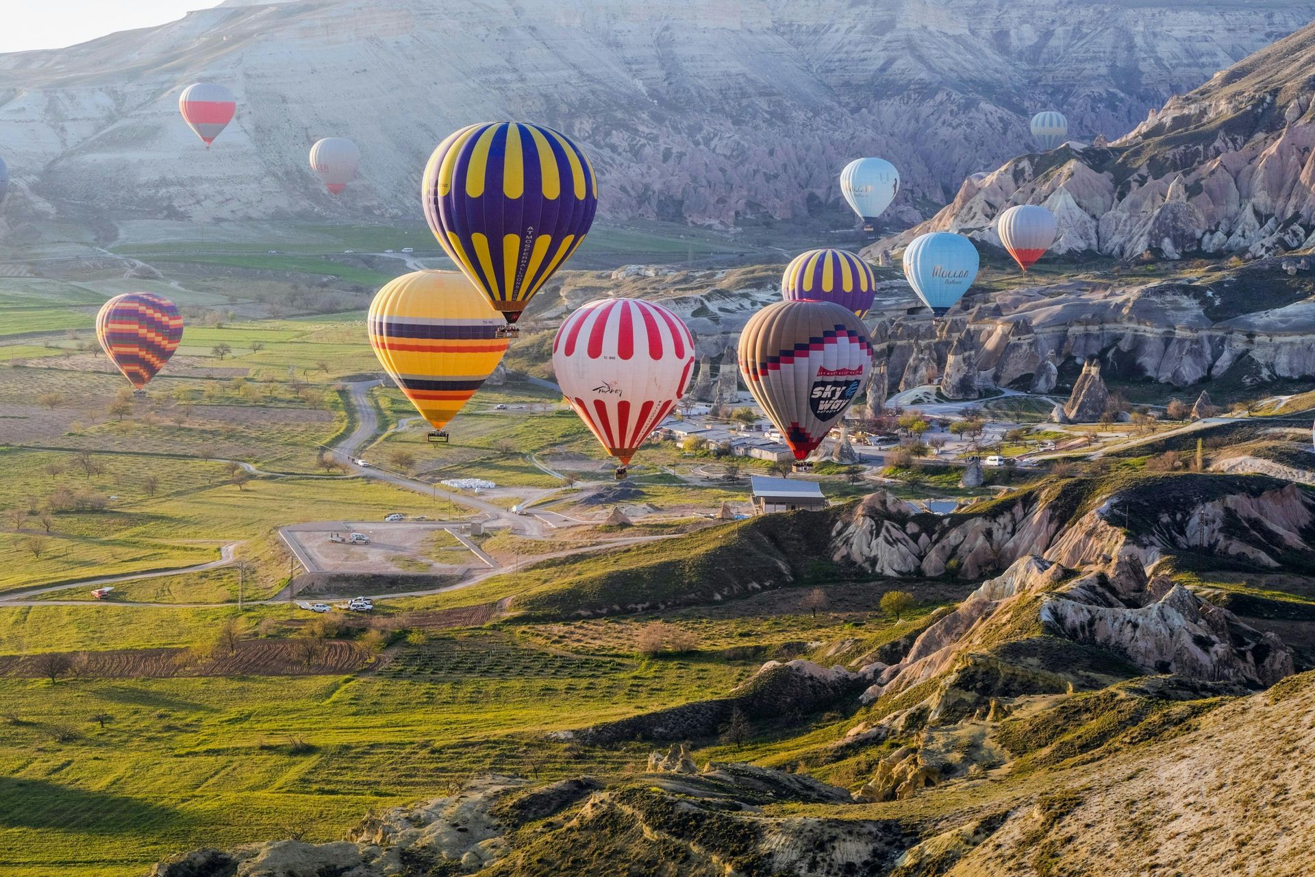 Hot air balloons over a green valley and rock formations in Cappadocia, Turkey, at sunrise.