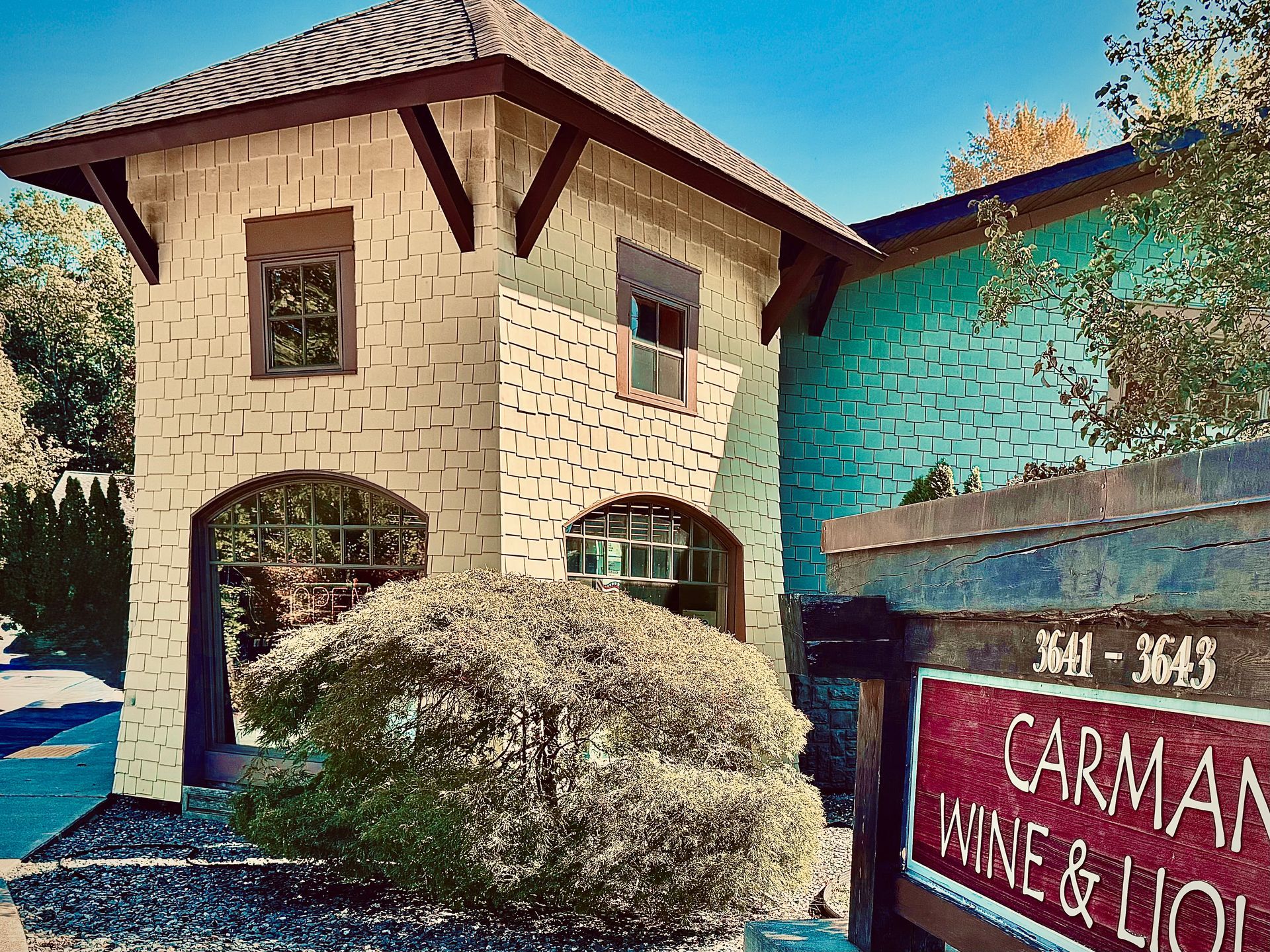 Carman Wine & Liquor store with a light-colored building, teal wall, and sign. A shrub is in the foreground.